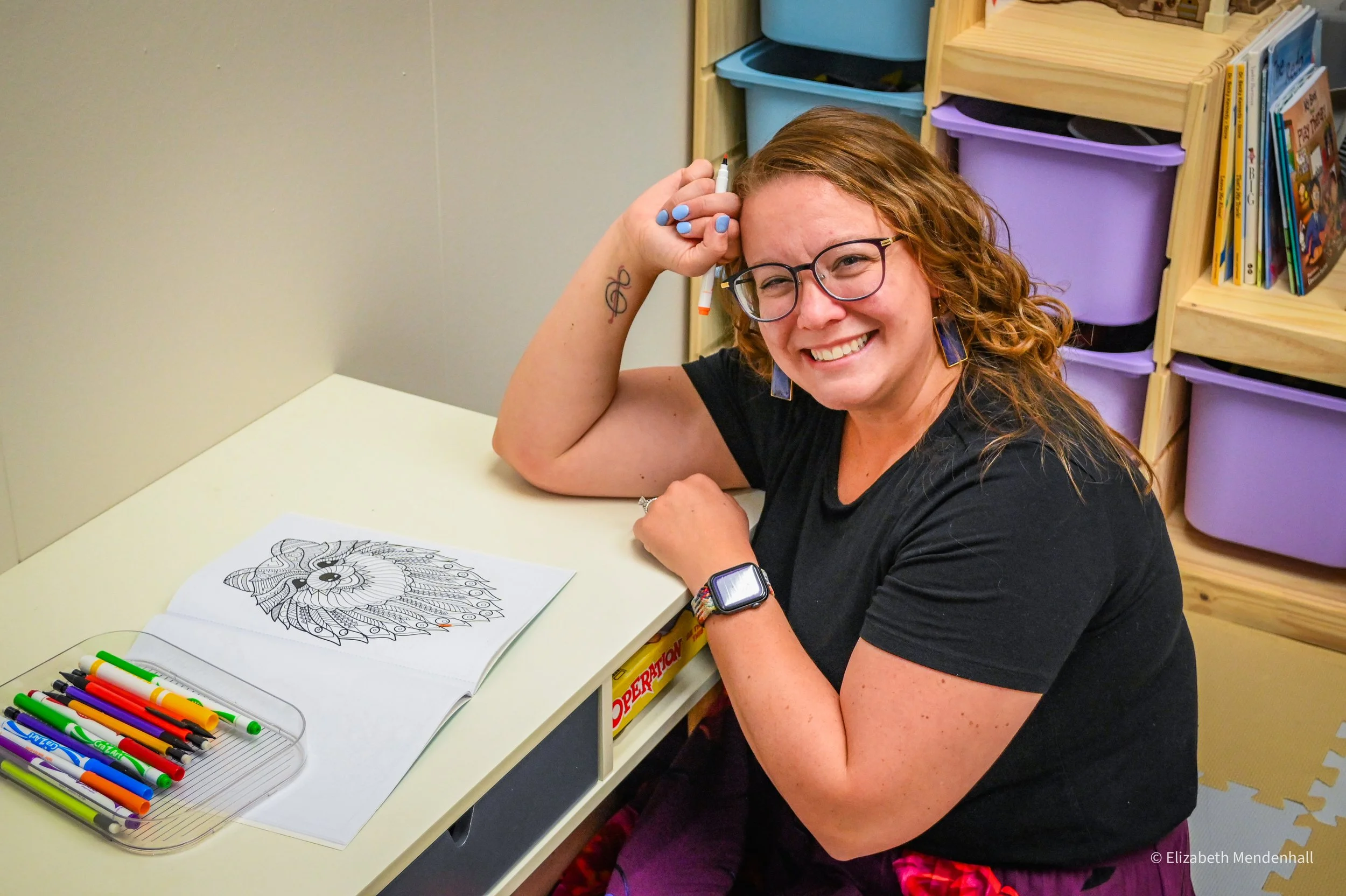 A woman with curly red hair and glasses is smiling and resting her head on her hand, sitting at a table in a classroom or library. She is wearing a black shirt and colorful accessories, and there is a coloring page with a detailed drawing of a bird or mythical creature in front of her. There are markers on the table, and colorful storage bins and bookshelves are visible in the background.