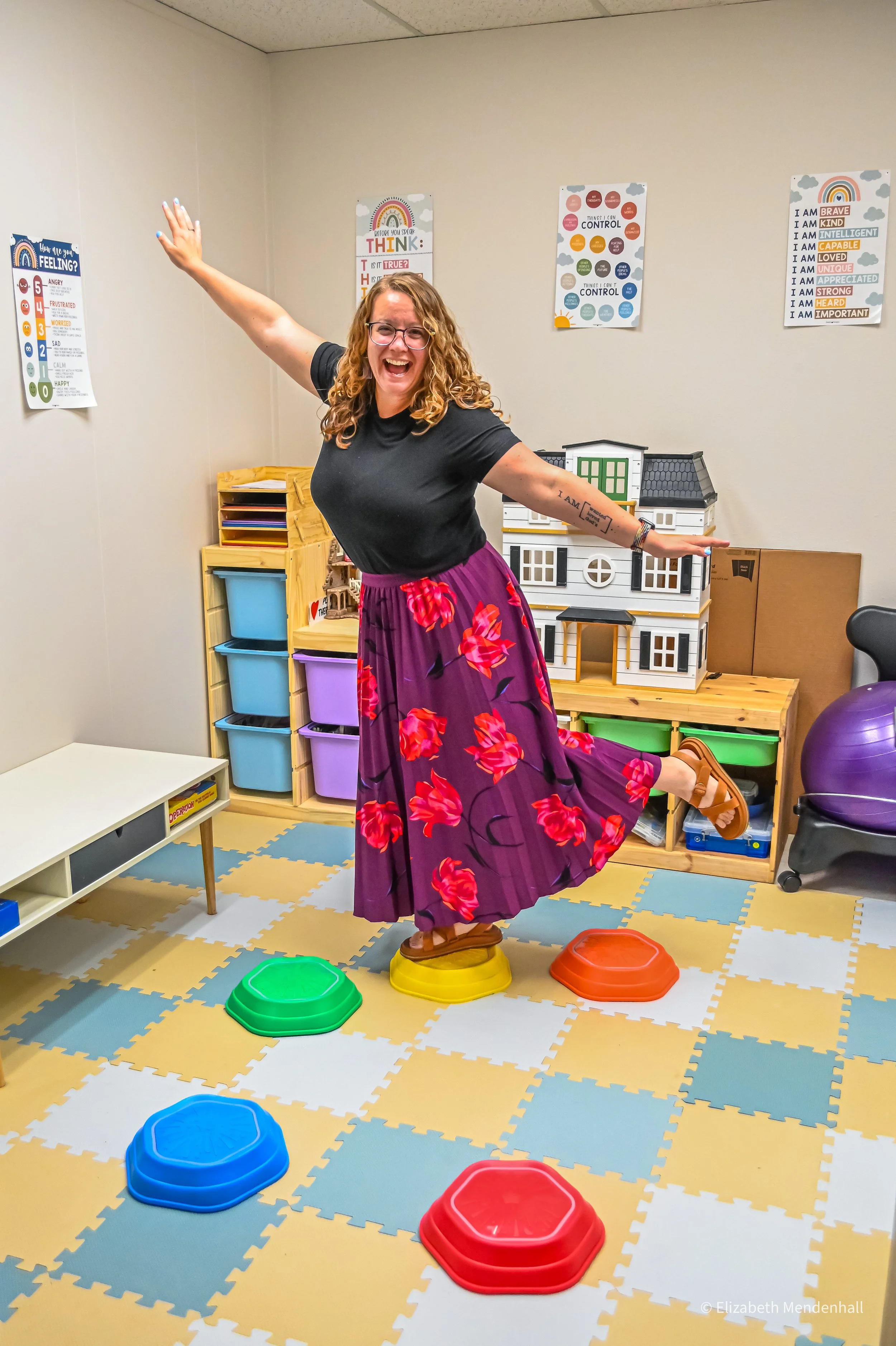 A woman with curly hair and glasses standing on a yellow stepping stone in a colorful playroom. She is smiling and raising her arms. The playroom has educational posters on the wall, a dollhouse, storage bins, and foam puzzle mats on the floor with blue, green, red, and yellow stepping stones.