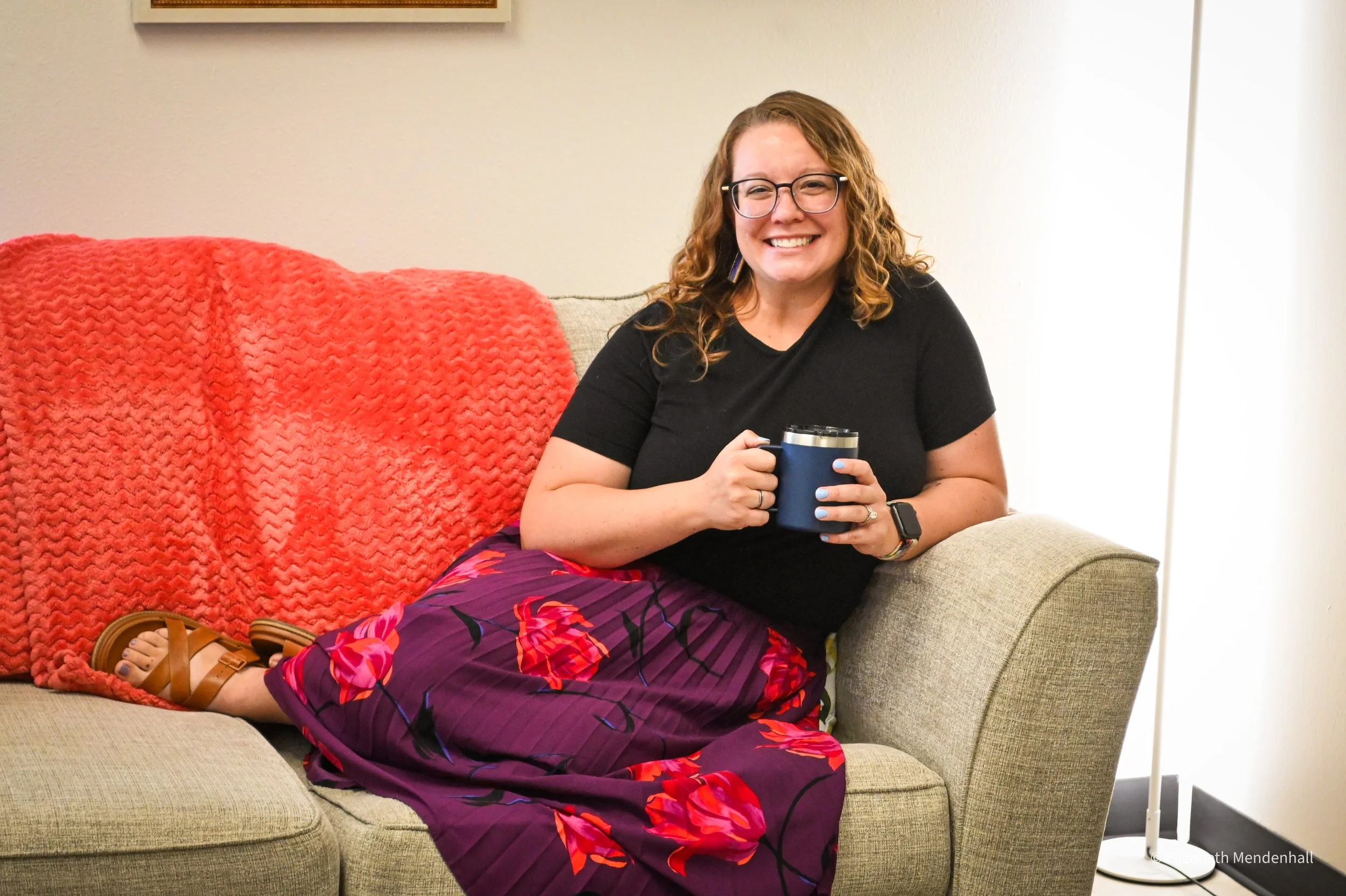 A woman with curly hair, glasses, and a black shirt sitting on a beige couch, smiling and holding a blue mug, with a red blanket on the back of the couch and purple floral skirt, inside a room with a white wall. The woman is Kerstin, the therapist