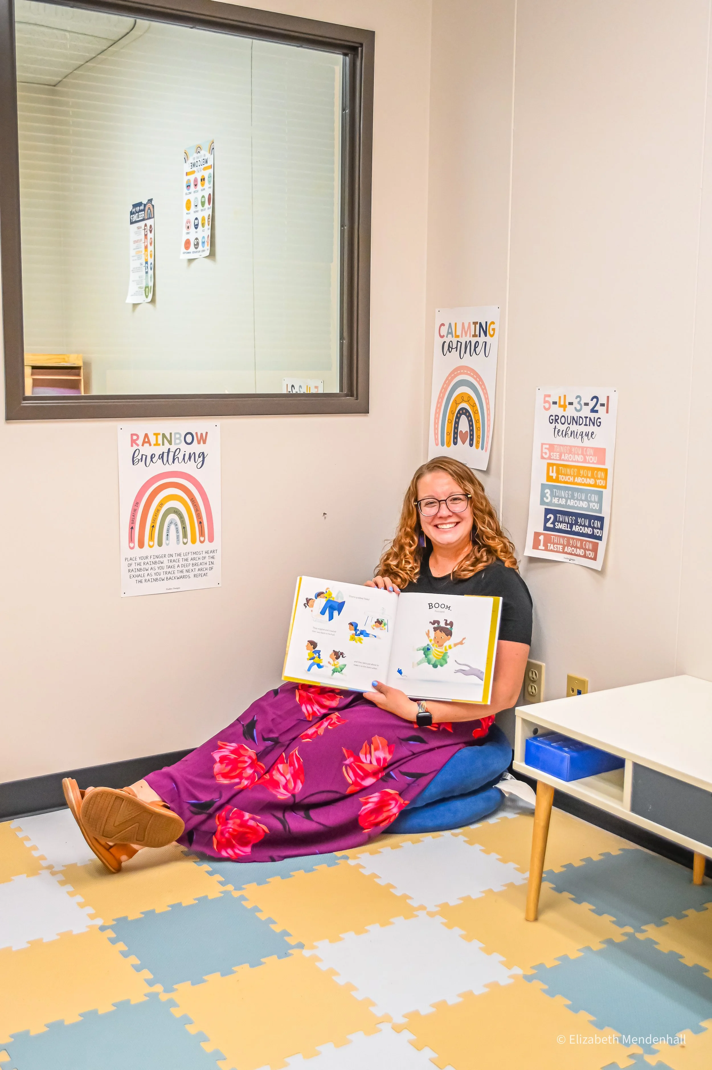 A woman with curly hair, glasses, and a watch sits on the floor wearing a purple skirt with red flowers, smiling while reading a children's book. She is in a room with calming wall posters that include a rainbow breathing guide and grounding techniques, and a colorful foam puzzle mat on the floor.