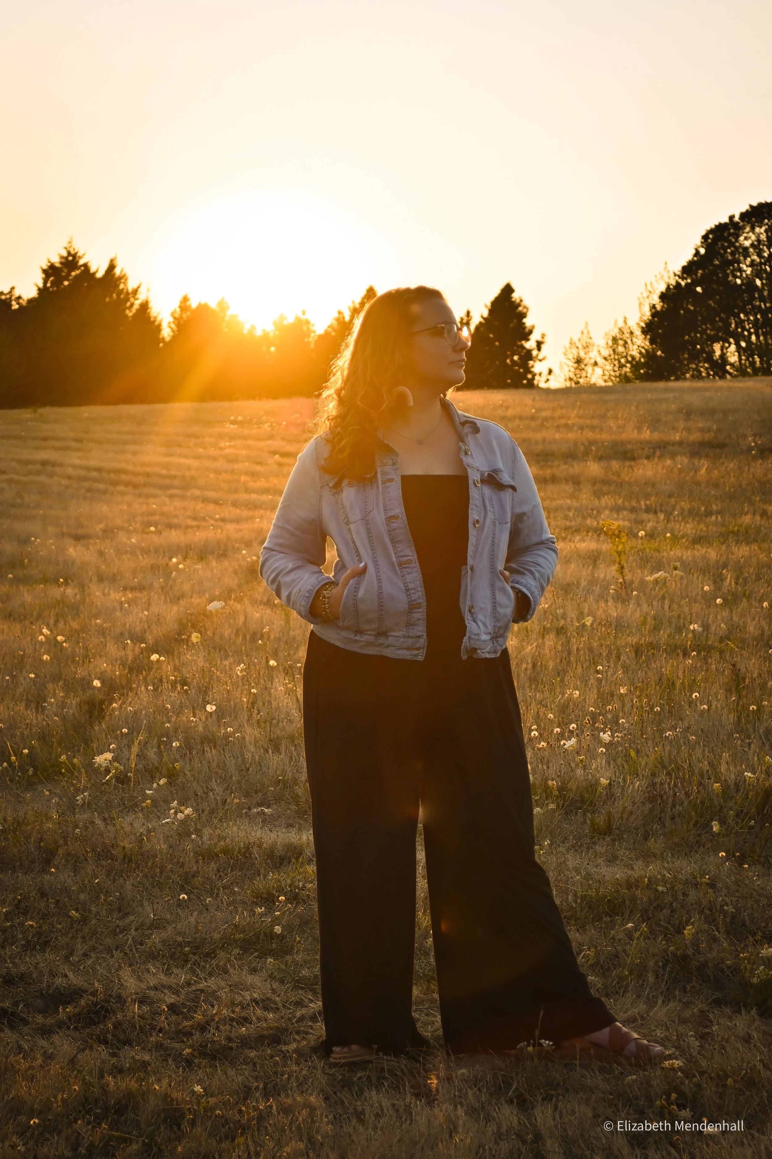 A young woman with glasses and curly hair stands in a field at sunset, wearing a denim jacket and black pants. The young woman is Kerstin, the therapist behind Limitless Growth Therapy.