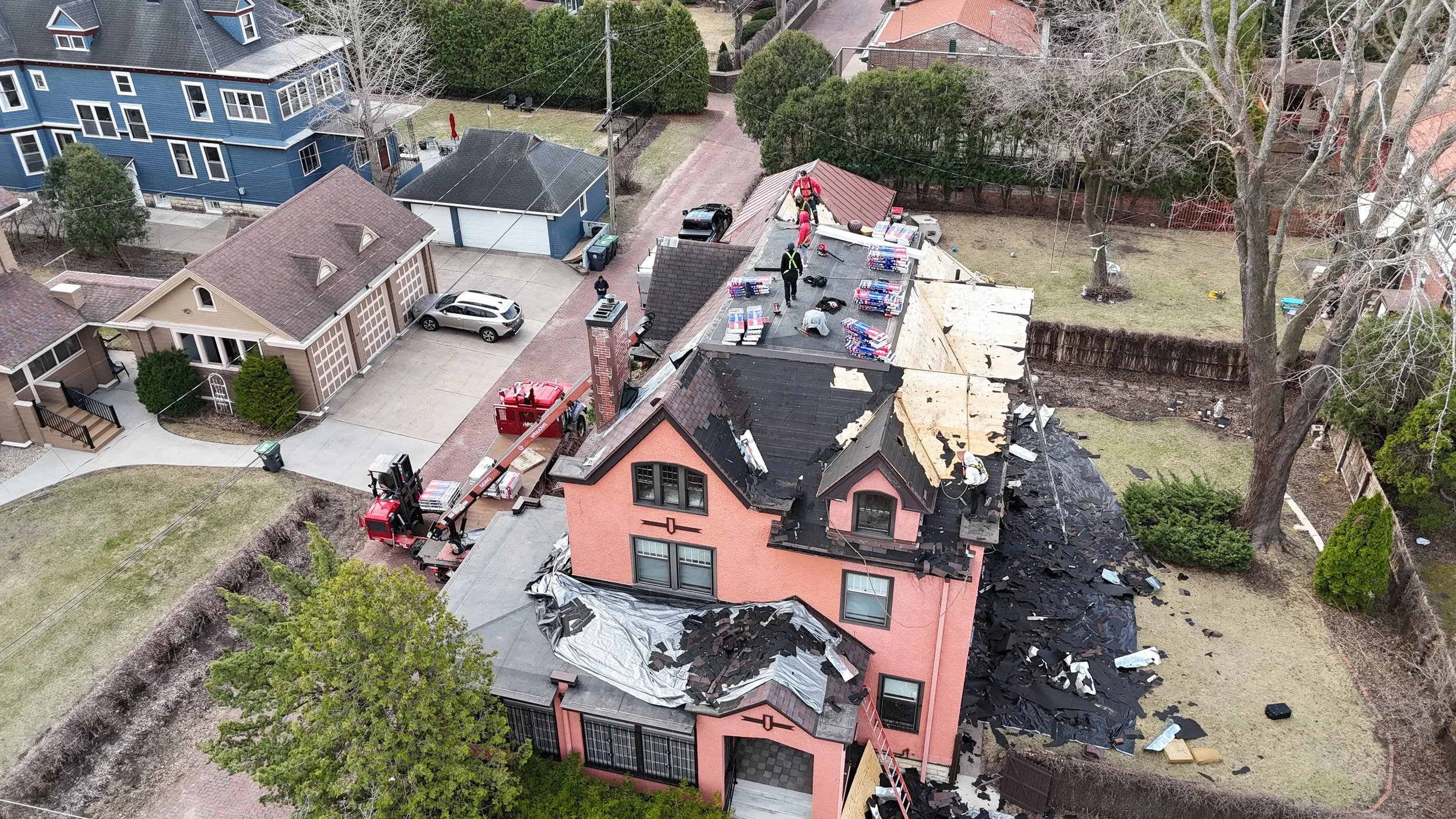 Aerial view of a house under renovation, with workers on the roof removing shingles and roofing materials. Construction equipment and materials are visible on the roof and in the yard.