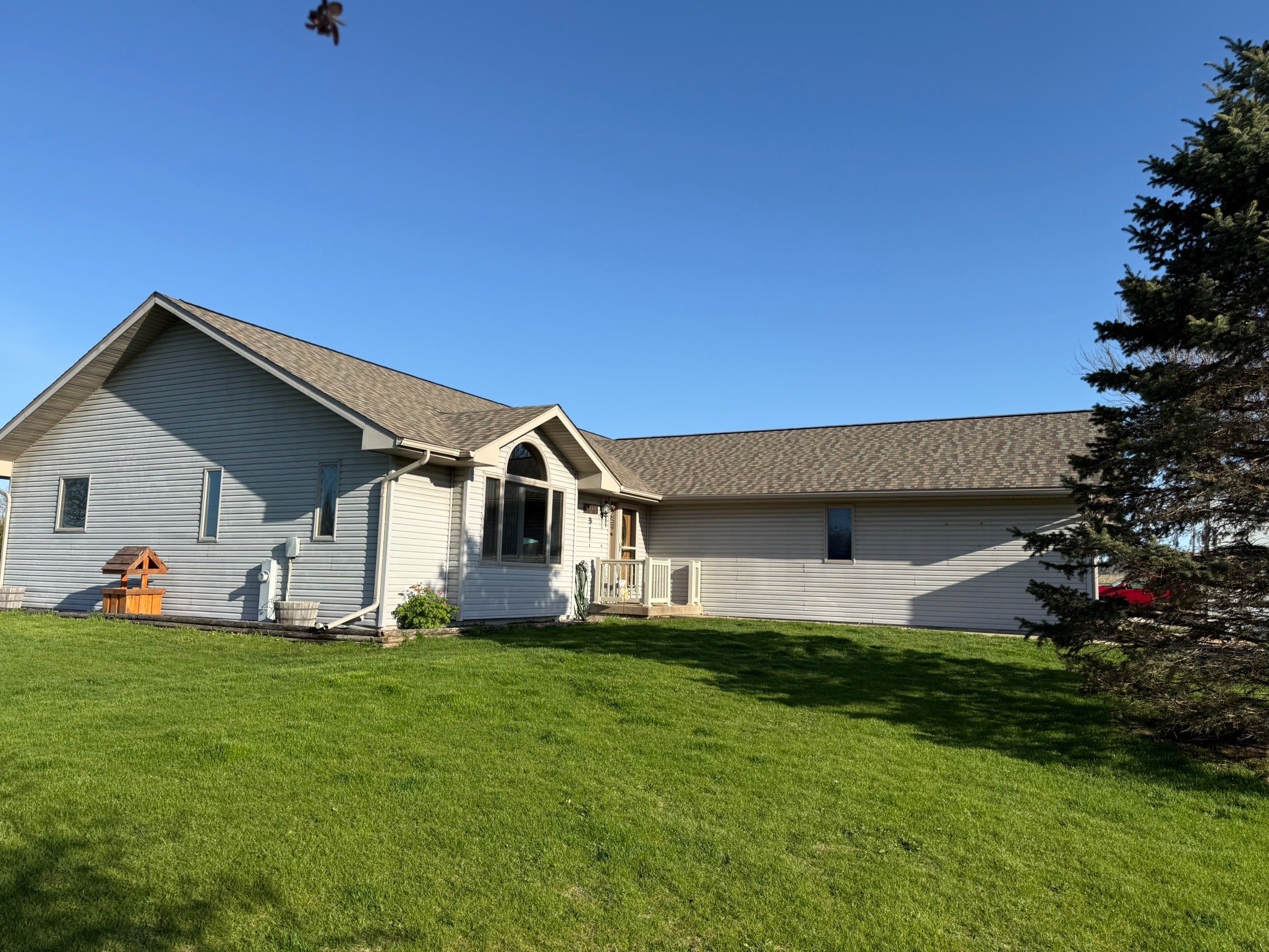 A light gray house with a steep roof and a large curved window, surrounded by a lush green lawn and a few trees, under a clear blue sky.