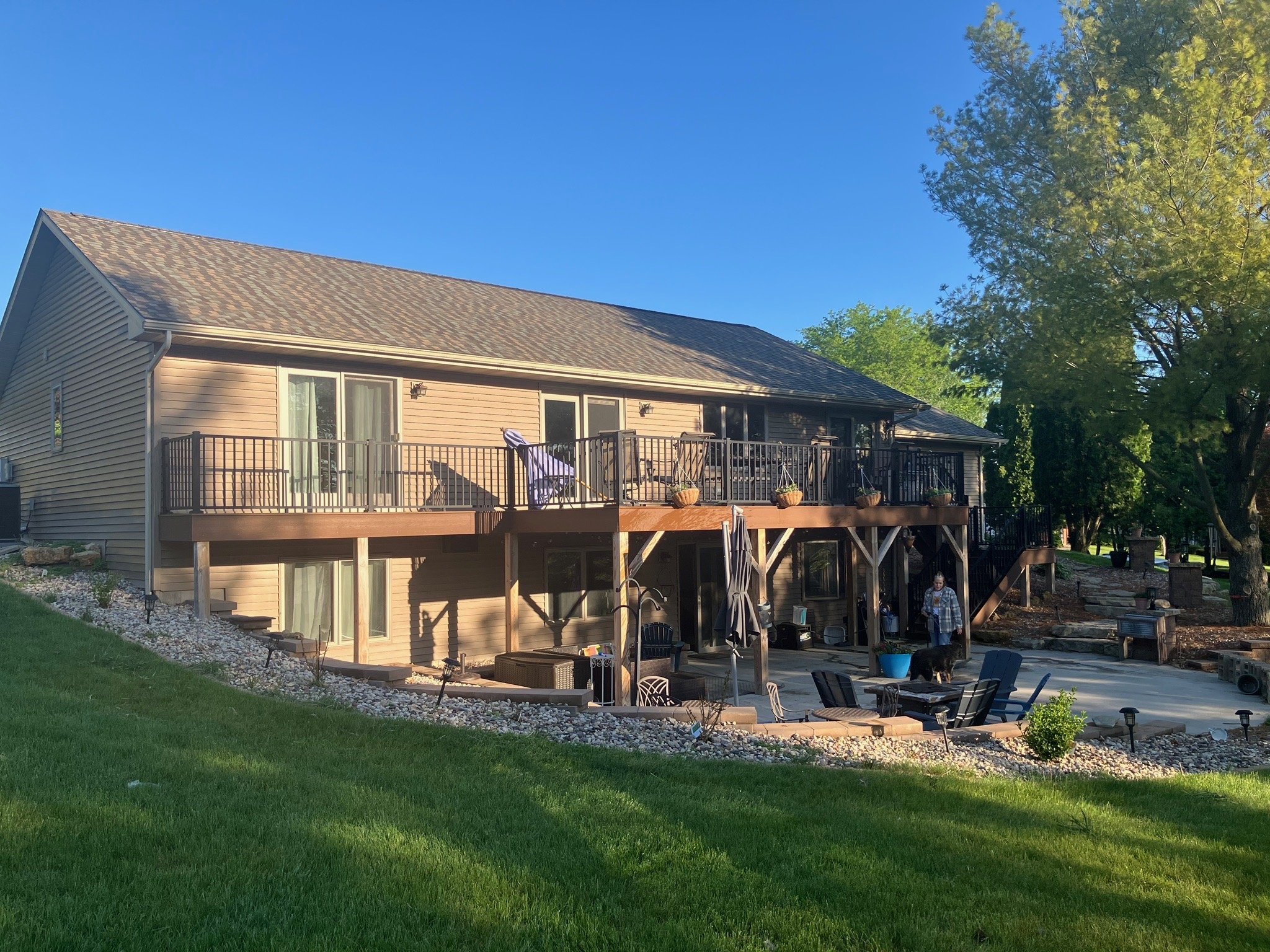 Backyard view of a two-story house with a wooden deck and patio, grassy lawn in foreground, trees, and clear blue sky.
