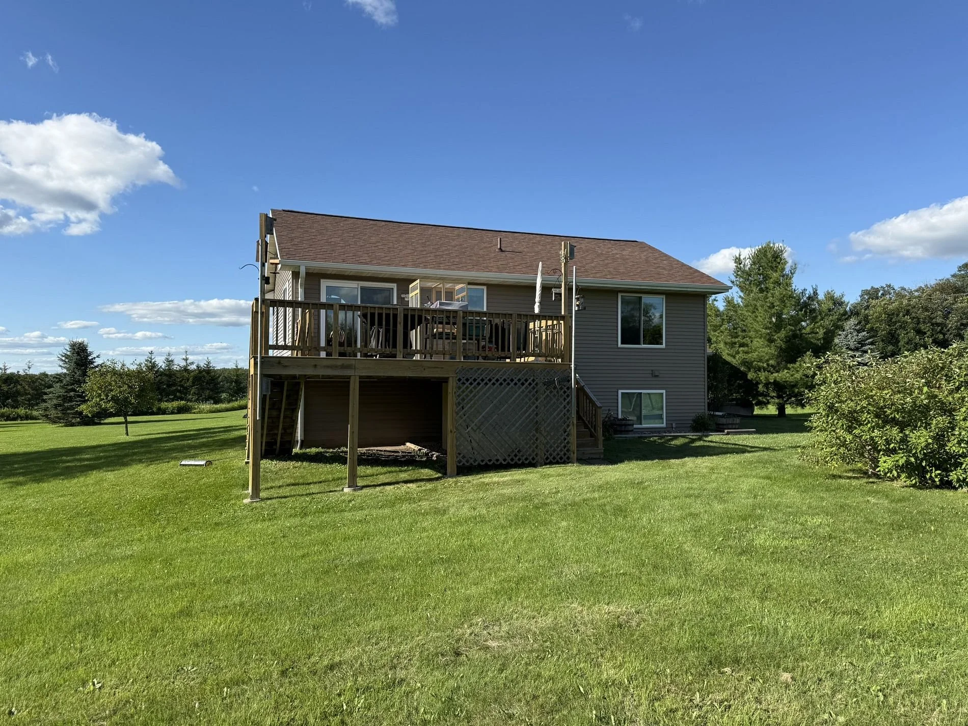 Backyard view of a two-story house with an elevated wooden deck and stairs, surrounded by a green lawn and trees under a clear blue sky.