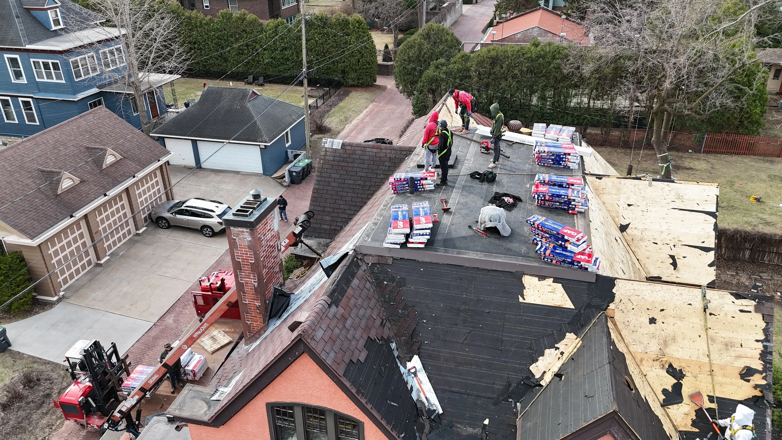 Aerial view of workers installing new roofing on a building with shingles, using equipment like a crane, on a residential street with parked cars, trees, and nearby houses.