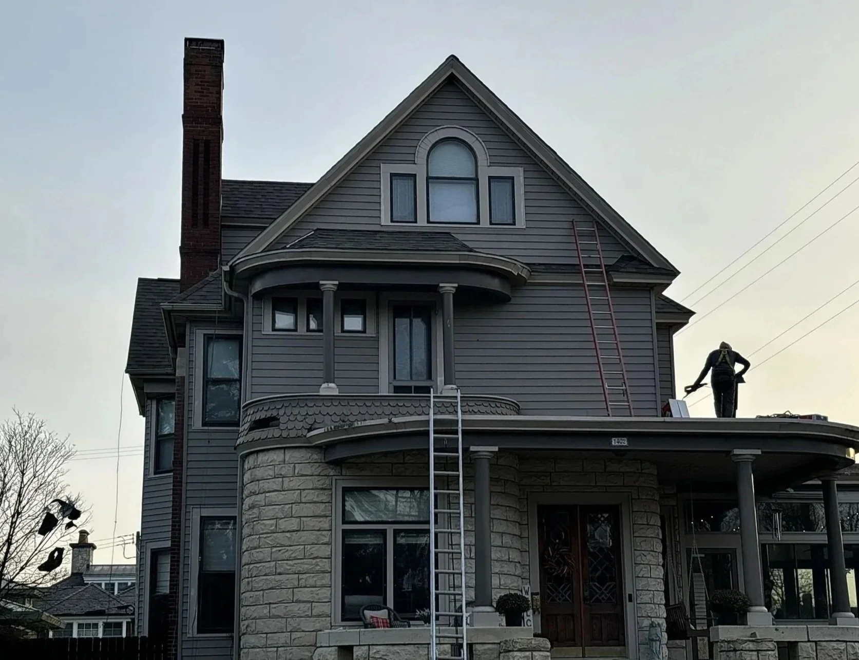 A large, multi-story house with gray siding, stone foundation, and a prominent chimney. A worker is on the roof with ladders leaning against the house, possibly doing maintenance or repairs.