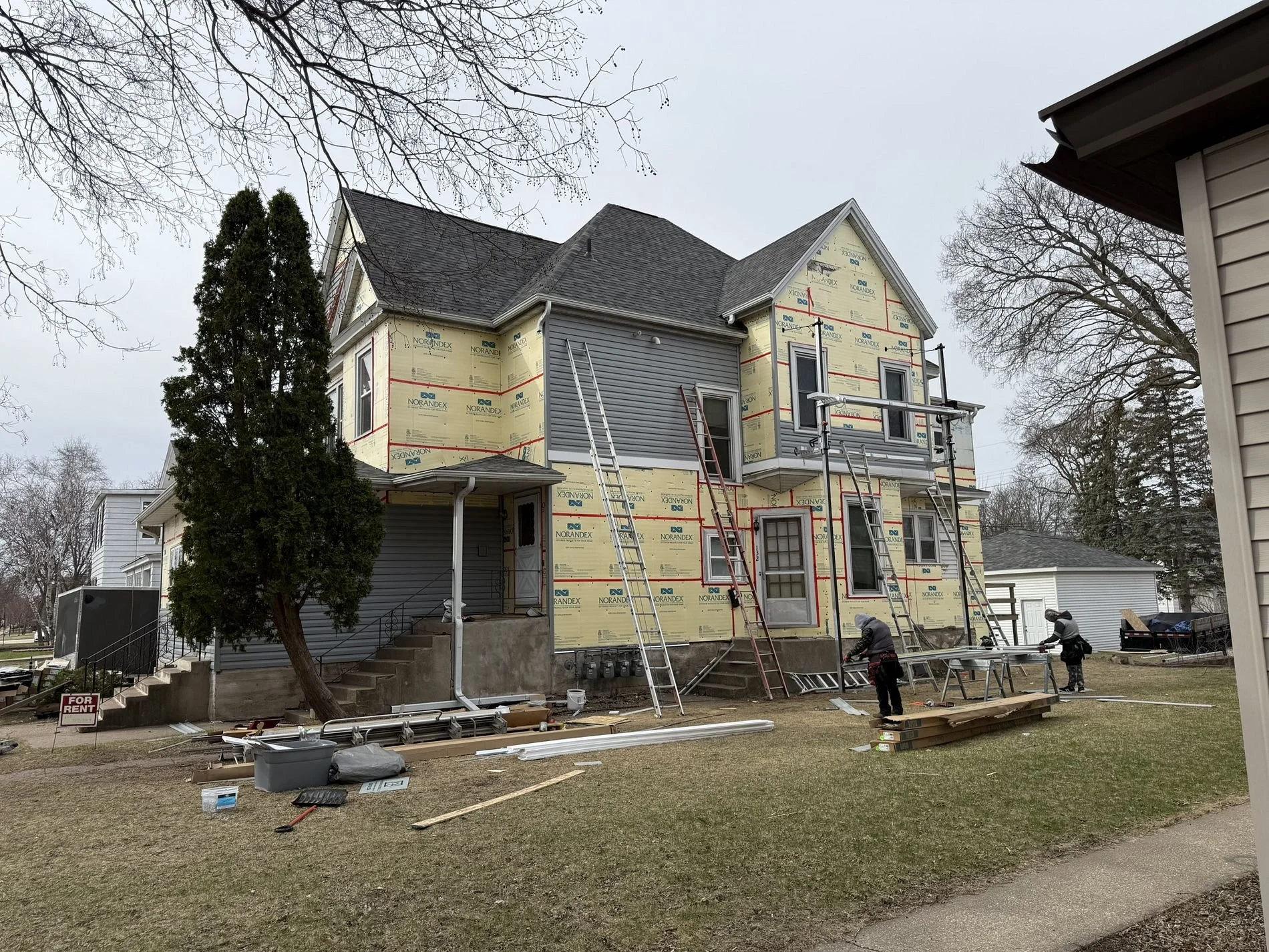 A house under construction with workers and scaffolding, partially covered in house wrap, and surrounded by building materials and equipment.