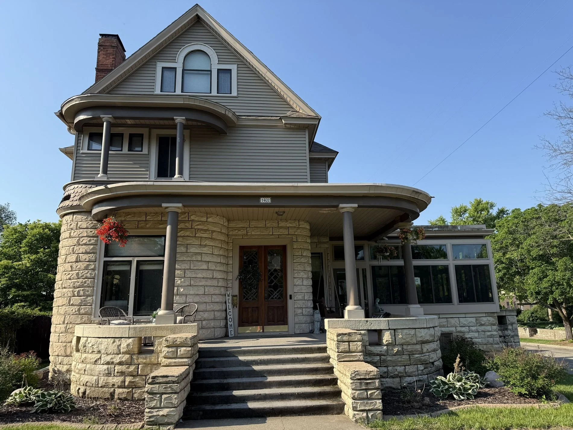 A large, multi-story house with a stone facade on the lower level and gray siding on the upper level, featuring a front porch with stone steps and columns, and a glass-enclosed sunroom on the right side. The house is surrounded by greenery and has hanging flower baskets.