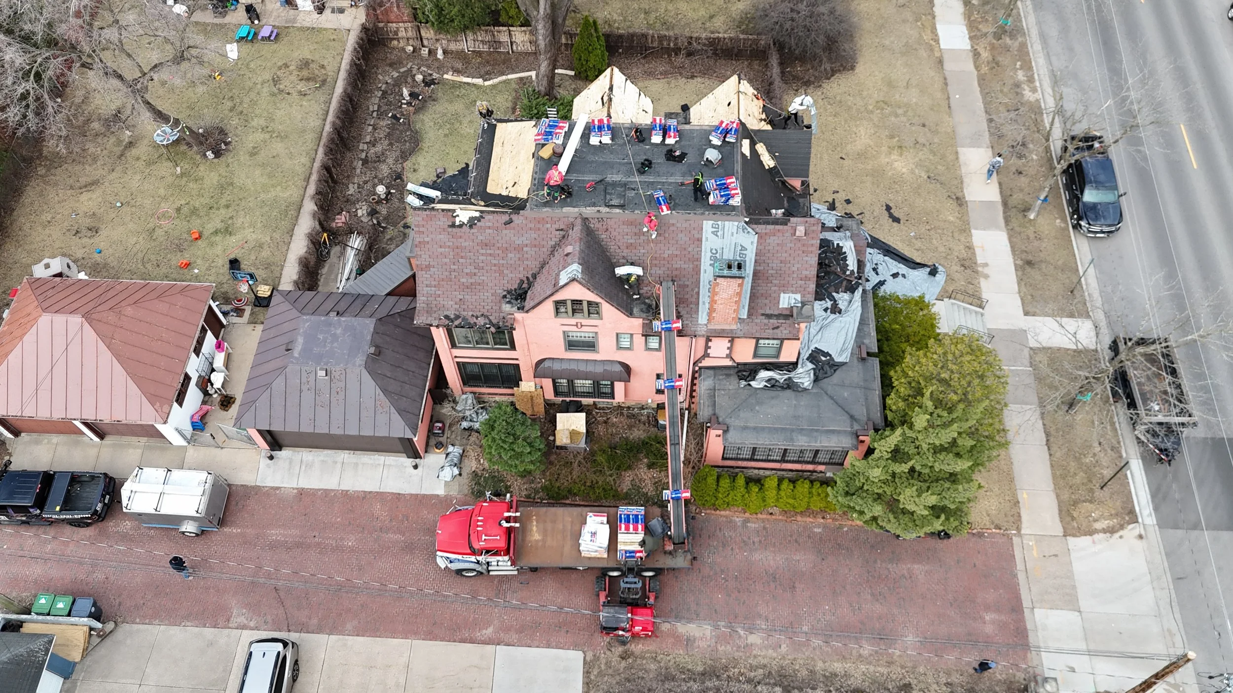 Aerial view of a house undergoing roof replacement, with workers, materials, and a crane on-site, surrounded by neighboring homes, trees, and a street with parked cars.
