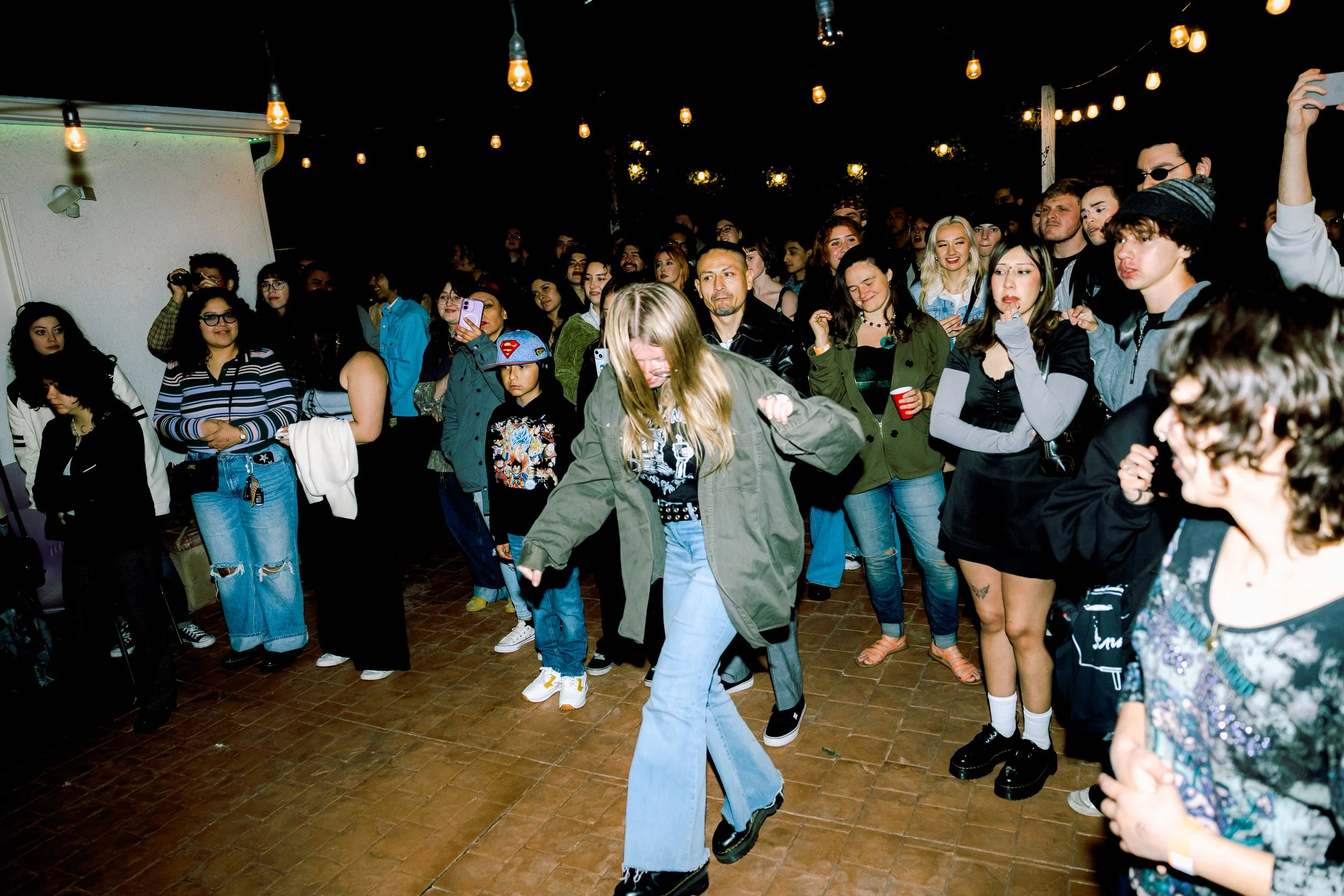 A group of people gathered outdoors at night, some watching and others dancing on a wooden floor, with string lights overhead.