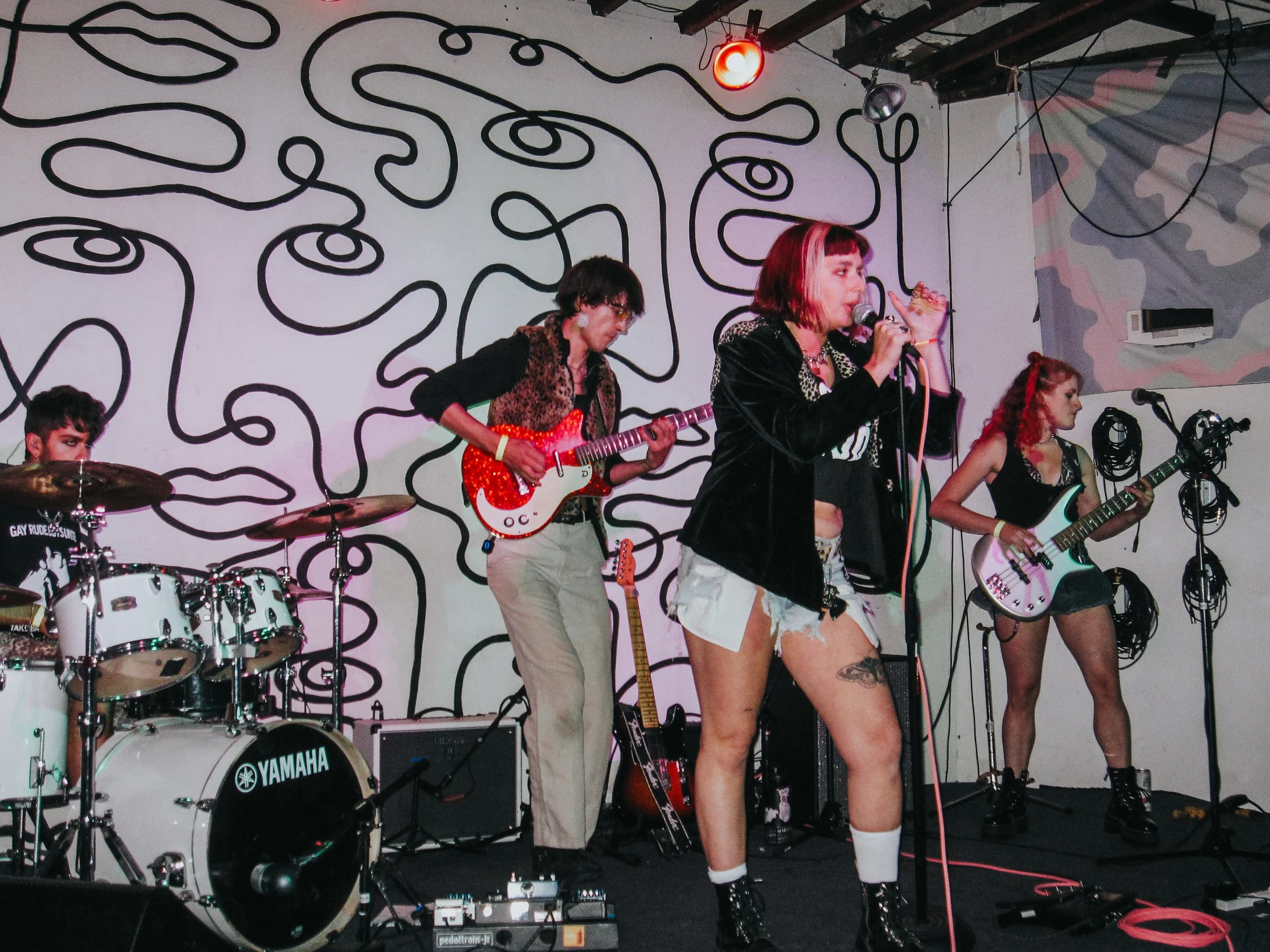 A band performing live on stage with a woman singing into a microphone, a guitarist, a bassist, and a drummer, in a room with abstract black line art on the wall.