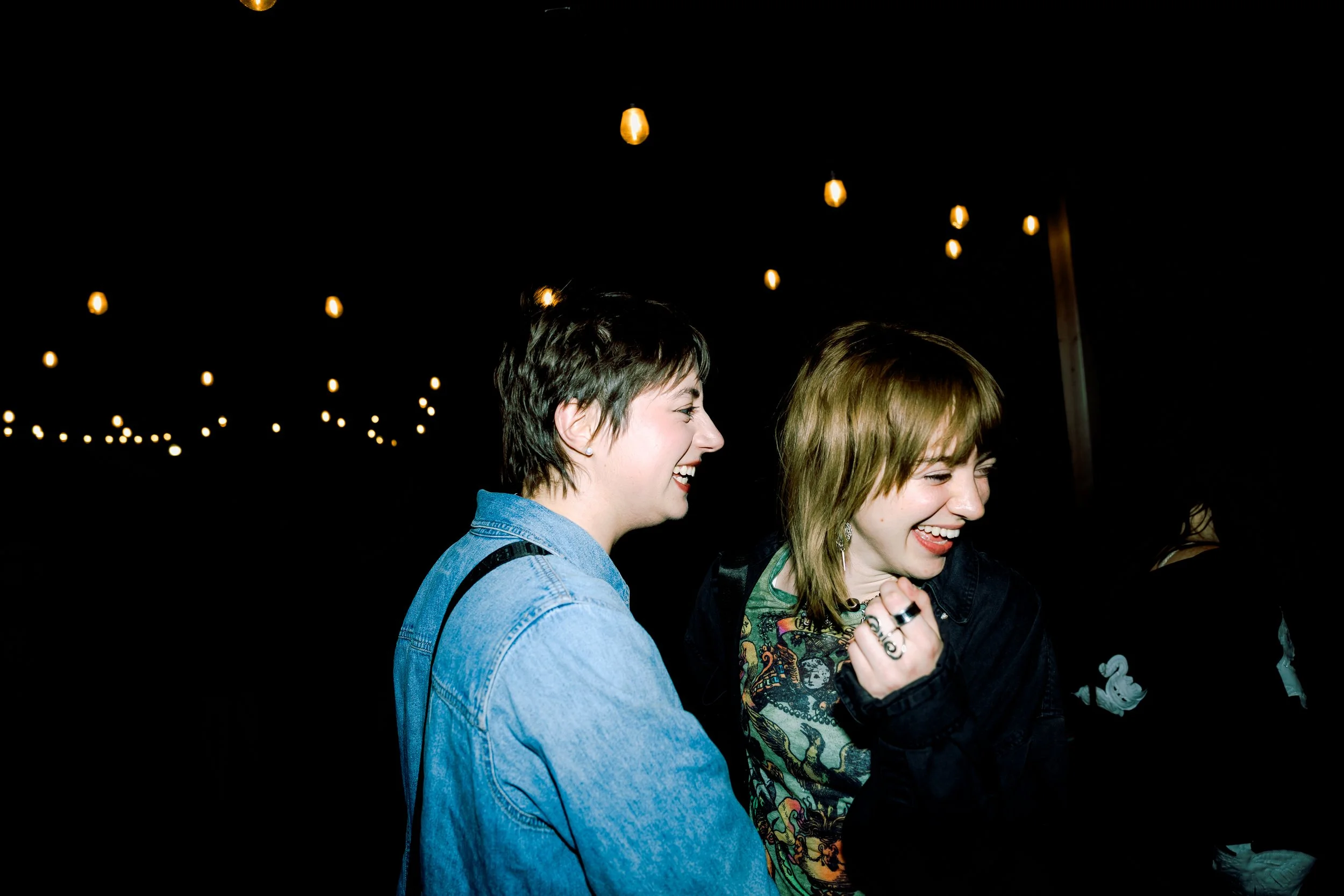 Two people laughing and enjoying a moment together at night, illuminated by string lights hanging above them.