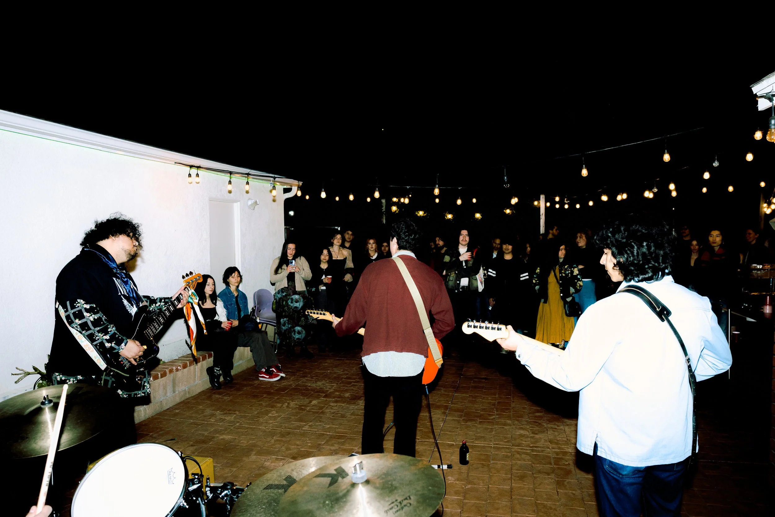 Musicians playing guitars and drums perform outdoors at night while an audience watches under string lights.
