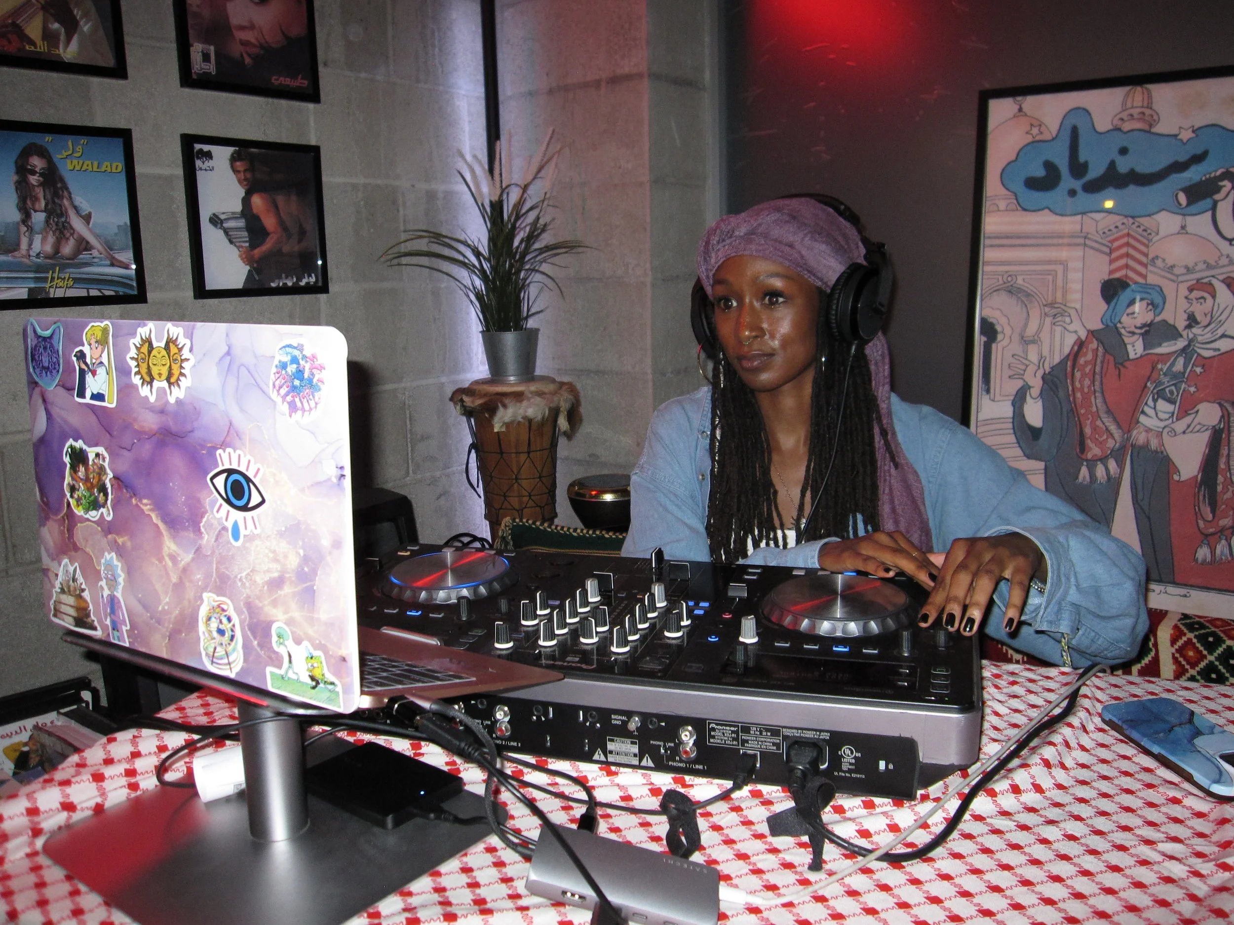 A woman DJ wearing a purple headscarf and headphones, operating a DJ controller with a colorful laptop on a red and white checkered table, in a room decorated with posters and wall art.