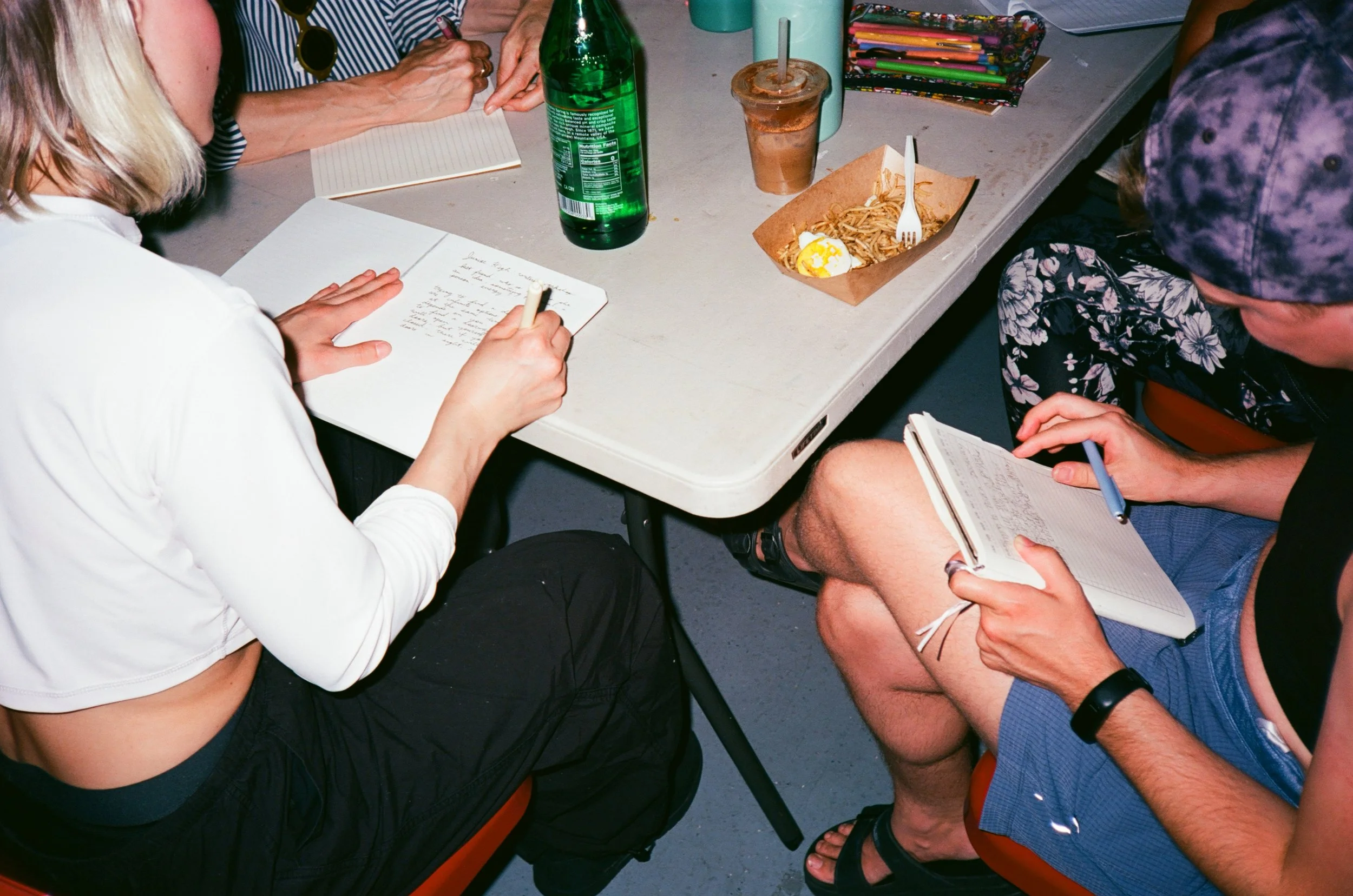 People sitting at a table with notebooks, drinks, and food, including a bottle of soda, an iced coffee, and a plate of noodles with a fork. One person is writing in a notebook, while another is taking notes in a smaller notebook.