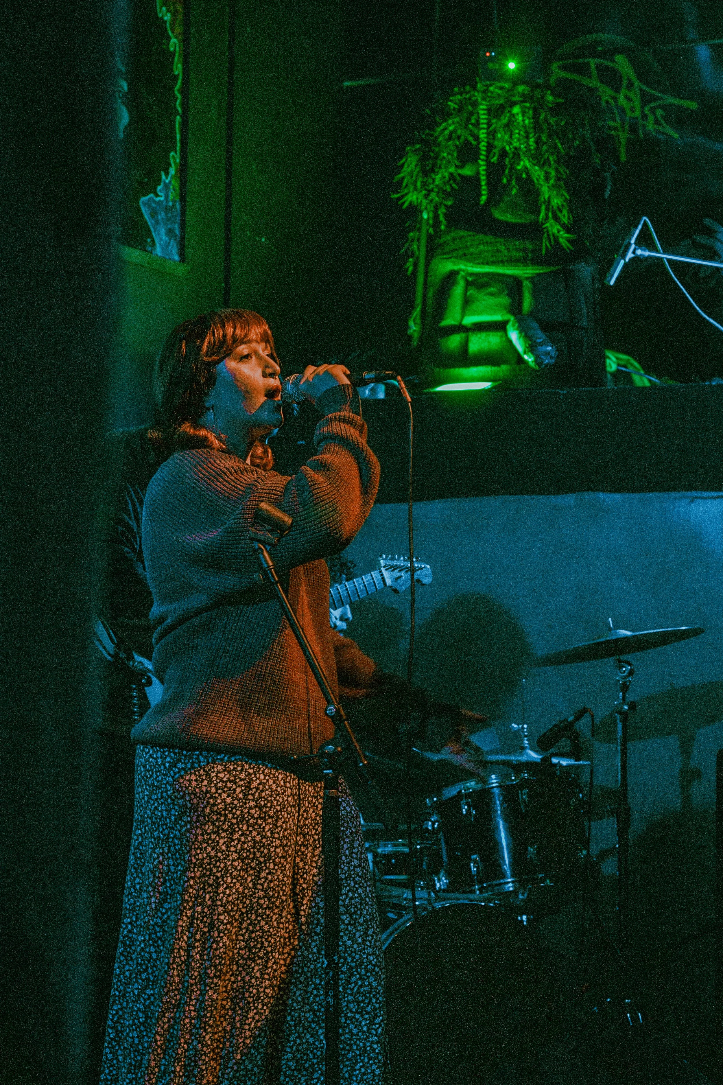 A woman singing into a microphone on a dimly lit stage with blue and green lighting, a drum set behind her, and a person playing guitar in the background.
