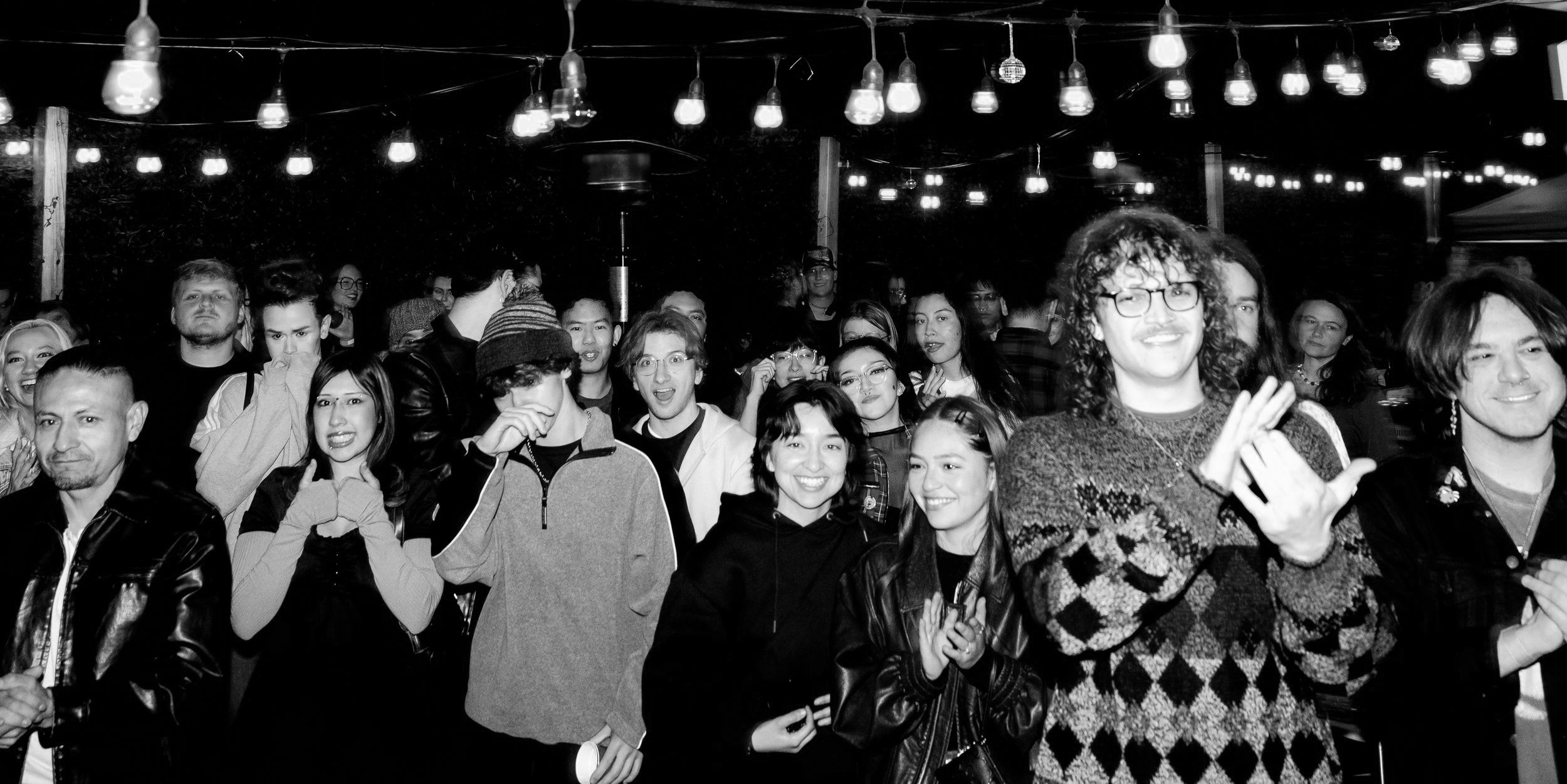 A group of people smiling and clapping at an outdoor night event, illuminated by string lights hanging overhead.