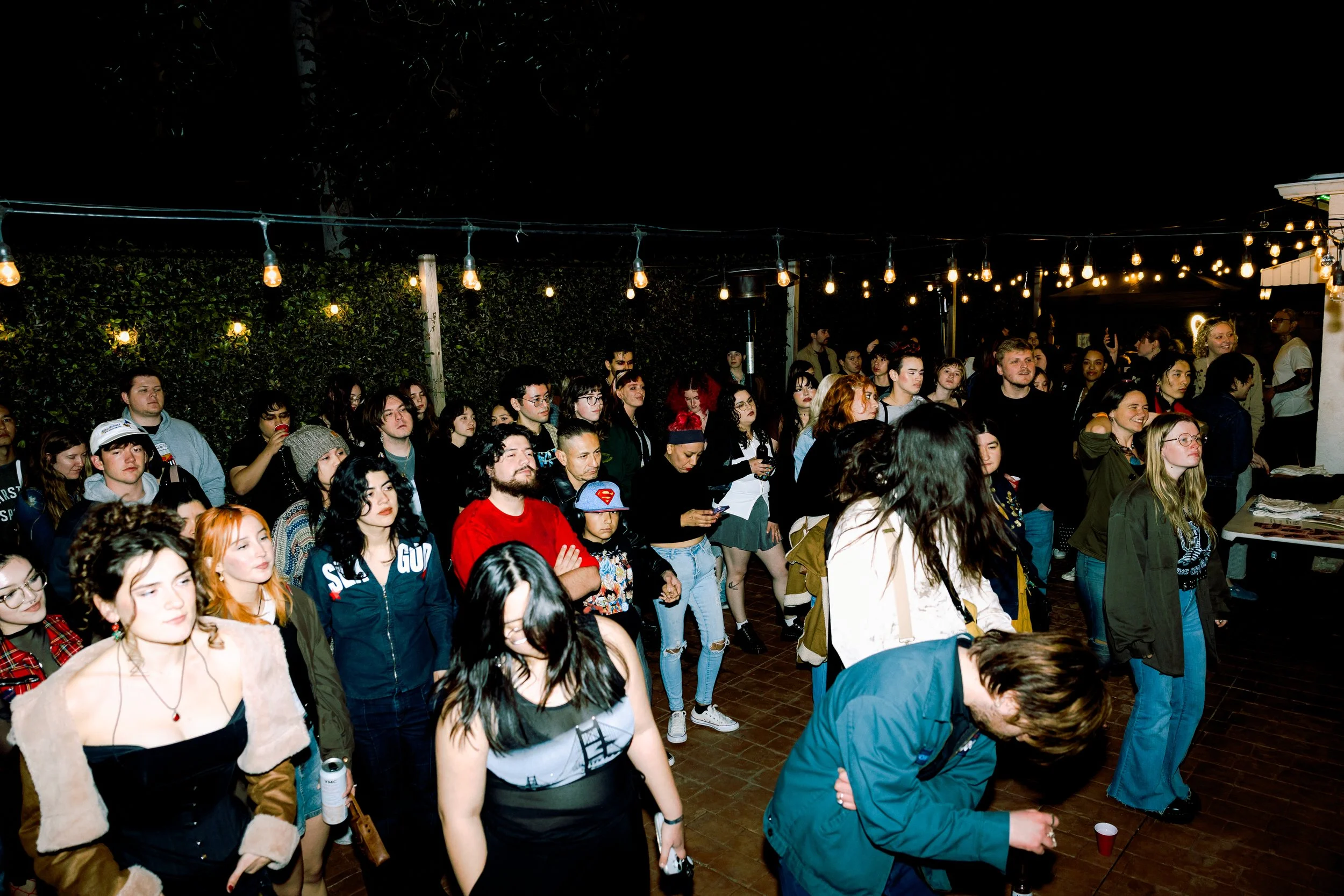 A crowd of people gathered at an outdoor event at night, under string lights, with some standing and some dancing.