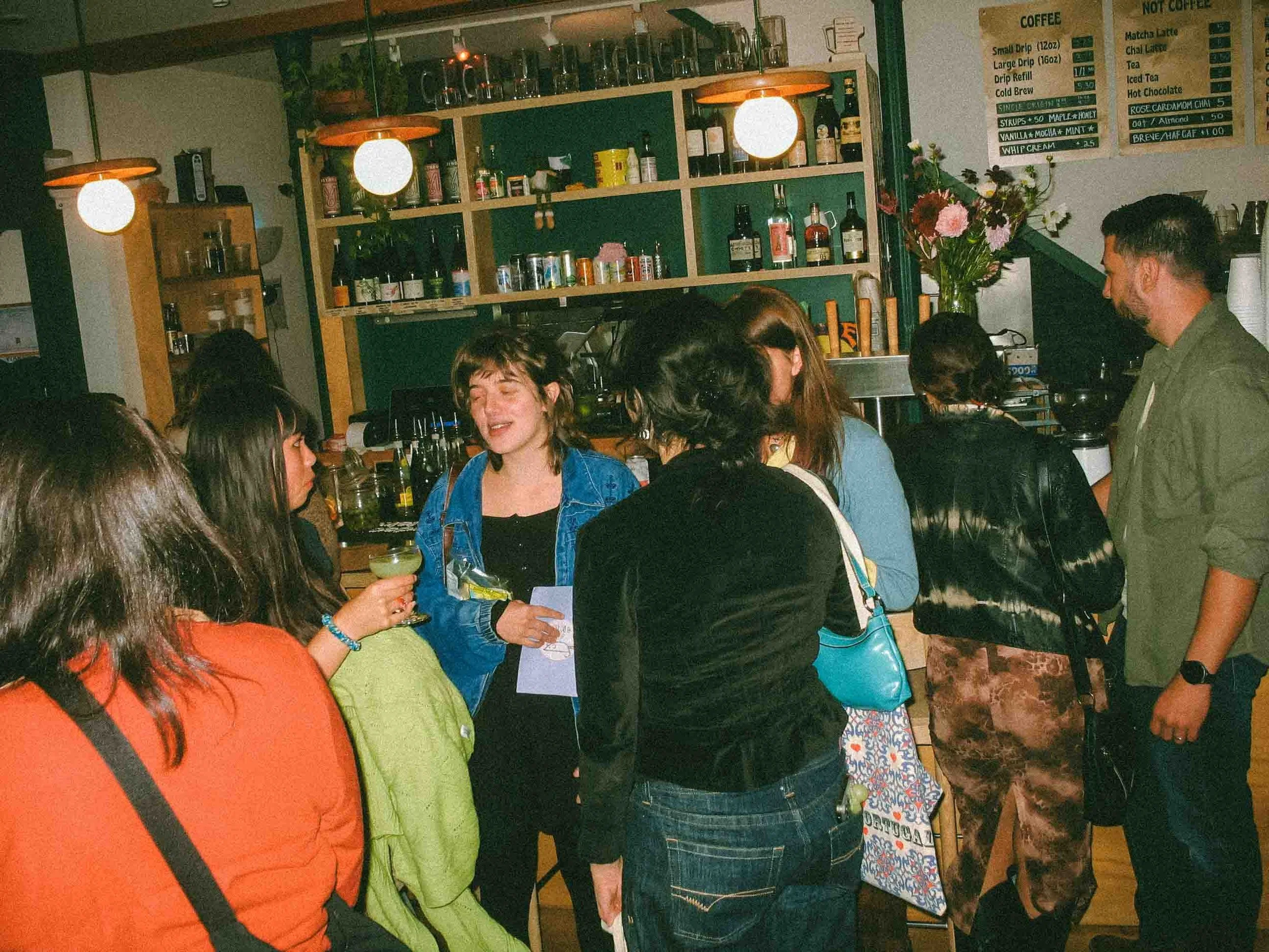A group of people gathered at a bar or cafe, engaging in conversation. The setting includes a shelf with bottles and supplies, and a menu board on the wall.