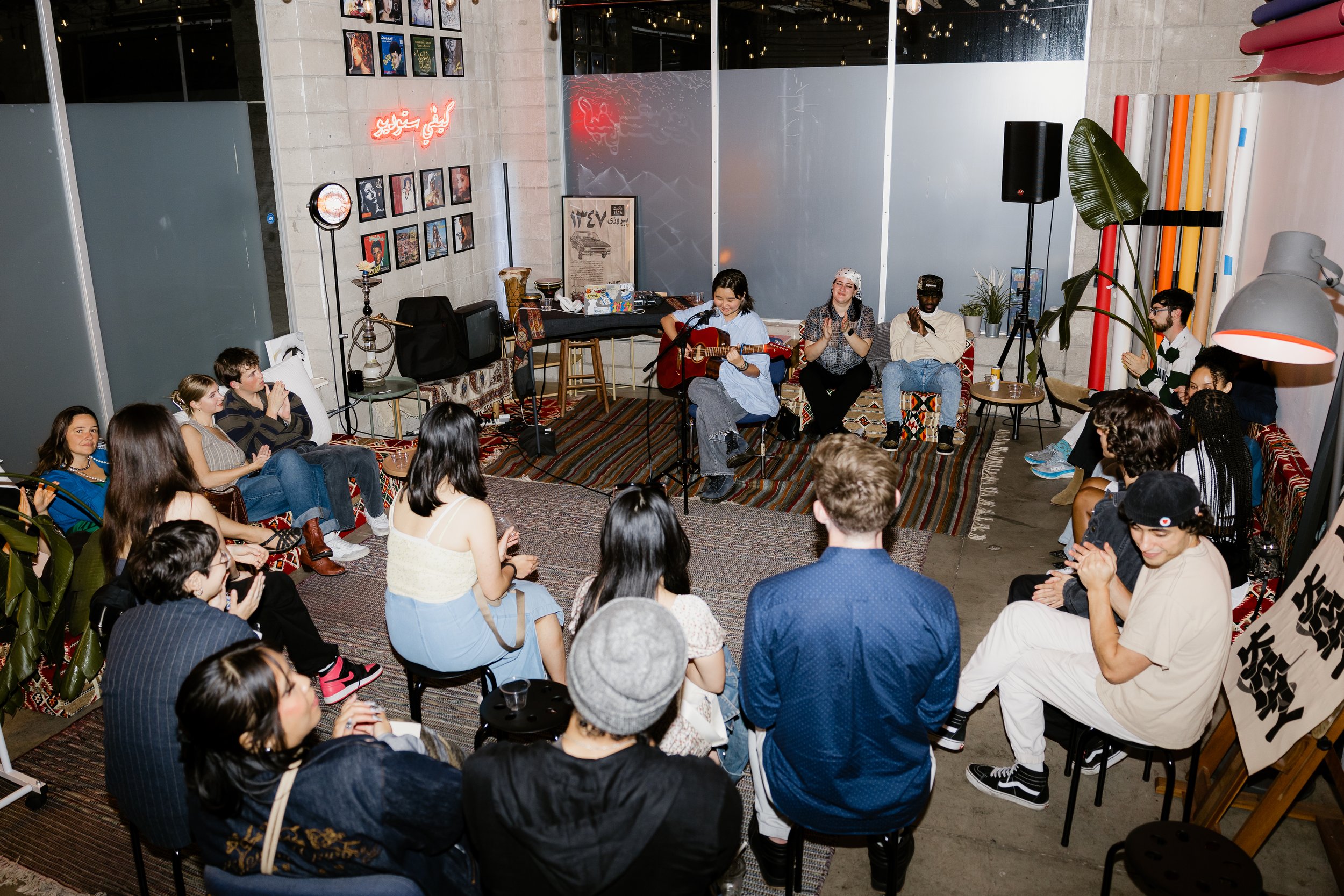 A group of people are gathered indoors, sitting in a semi-circle around a musician playing an acoustic guitar and singing. The setting has artwork on the wall, plants, and a casual, cozy atmosphere.