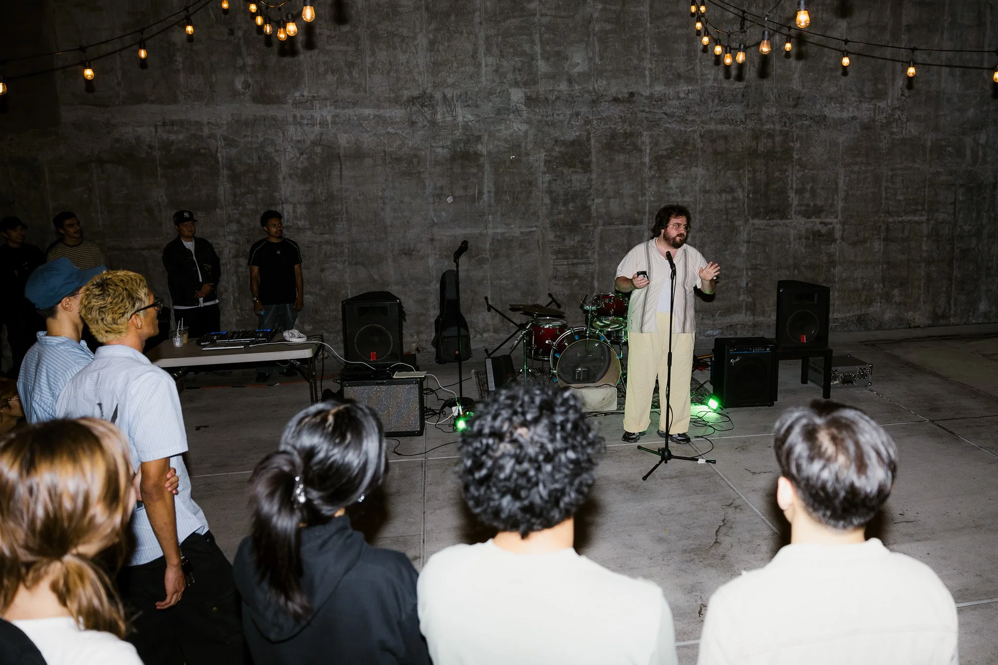 A man speaking on stage with a microphone in an indoor venue, with musical instruments set up behind him and string lights overhead, as an audience watches.