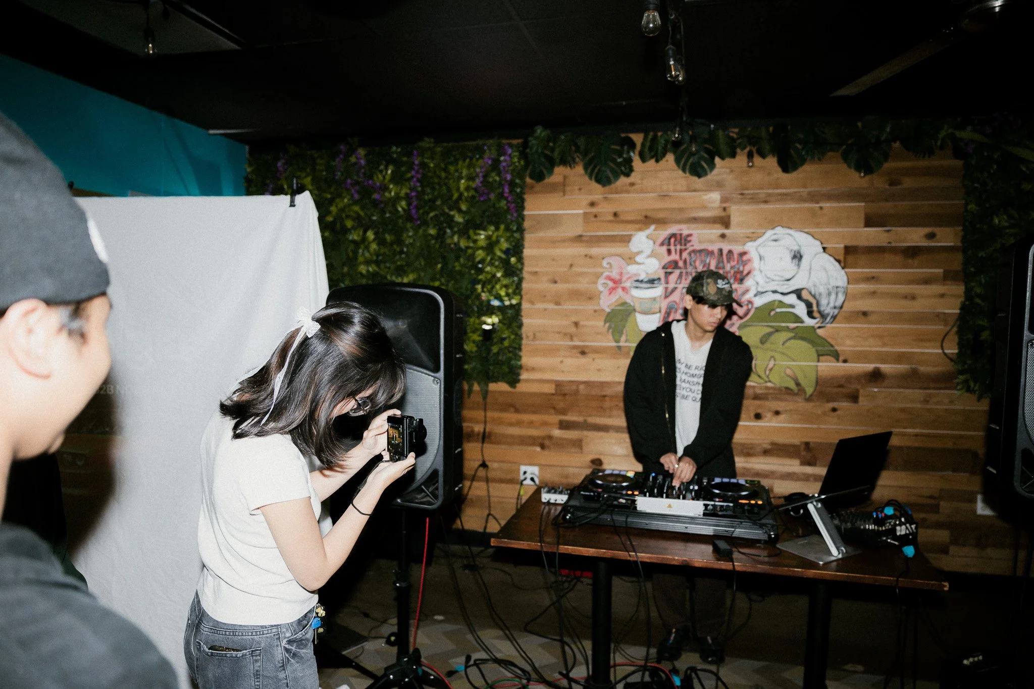 A DJ performing on a wooden stage with green plants and a mural in the background, while a young woman takes a photo and a person in the foreground watches.