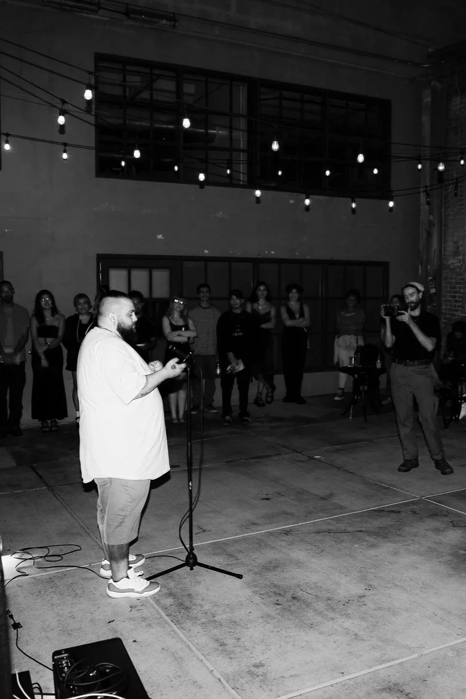 A man passionately speaking in front of a crowd at night in an outdoor urban setting, with string lights hanging overhead and people watching attentively.
