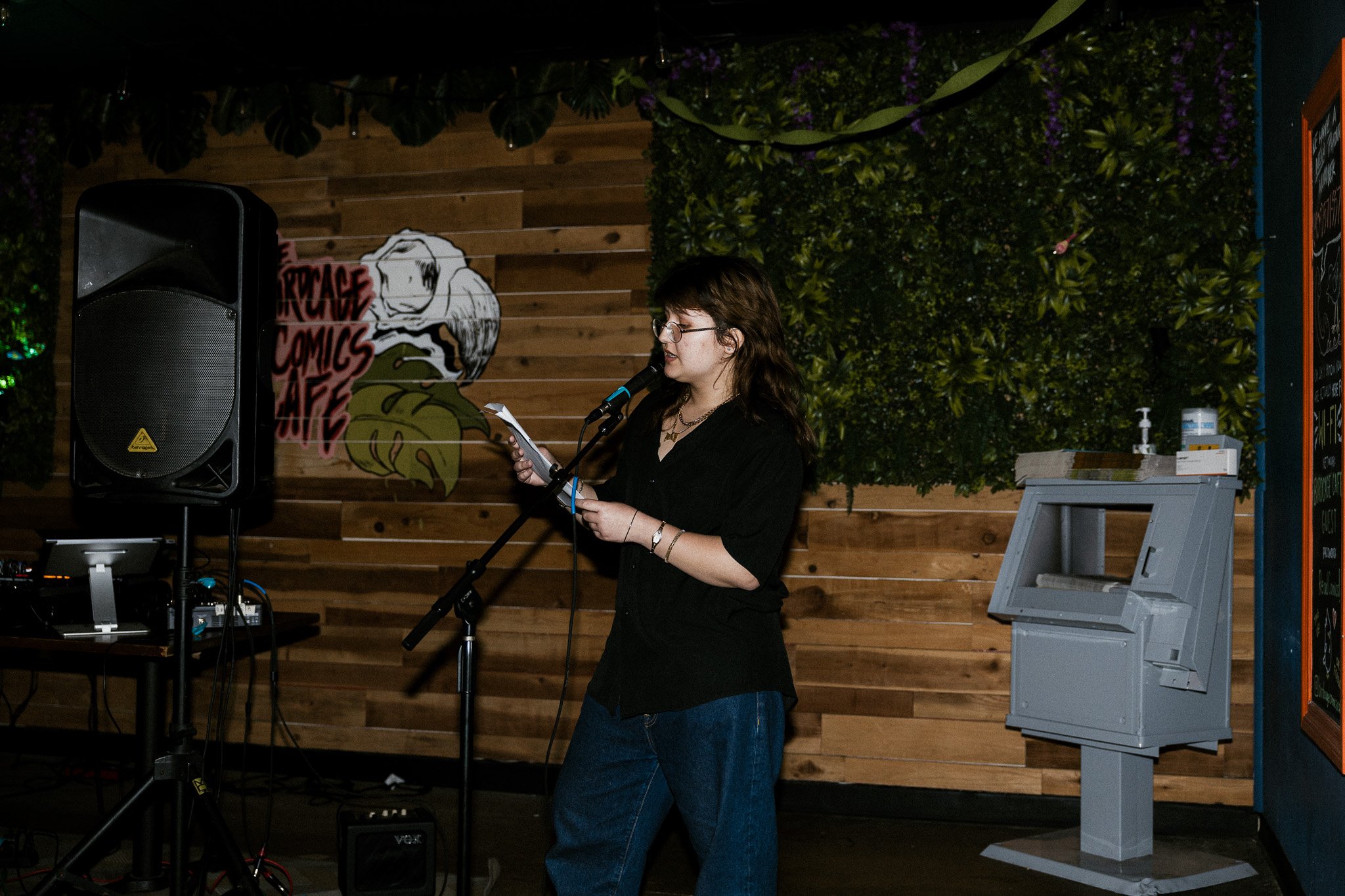 A woman with brown hair and glasses stands at a microphone, reading from a sheet of paper in a dimly lit venue with wooden walls and greenery. Behind her, a large speaker and a sign with a gorilla and text are visible. There is a gray piano or keyboa
