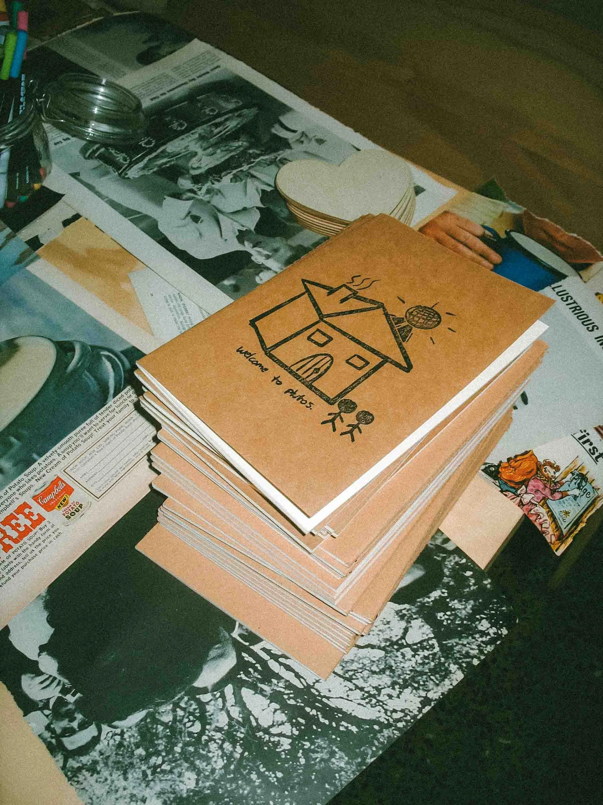 Stack of illustrated notebooks and zines on a cluttered desk.