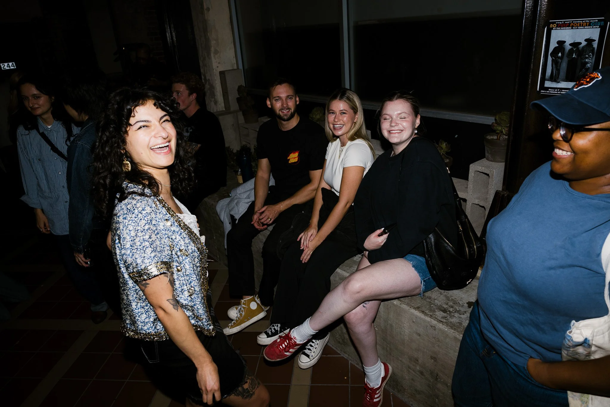 Group of people smiling and having fun at a social gathering, with some seated on a concrete bench and others standing in a casual indoor setting.