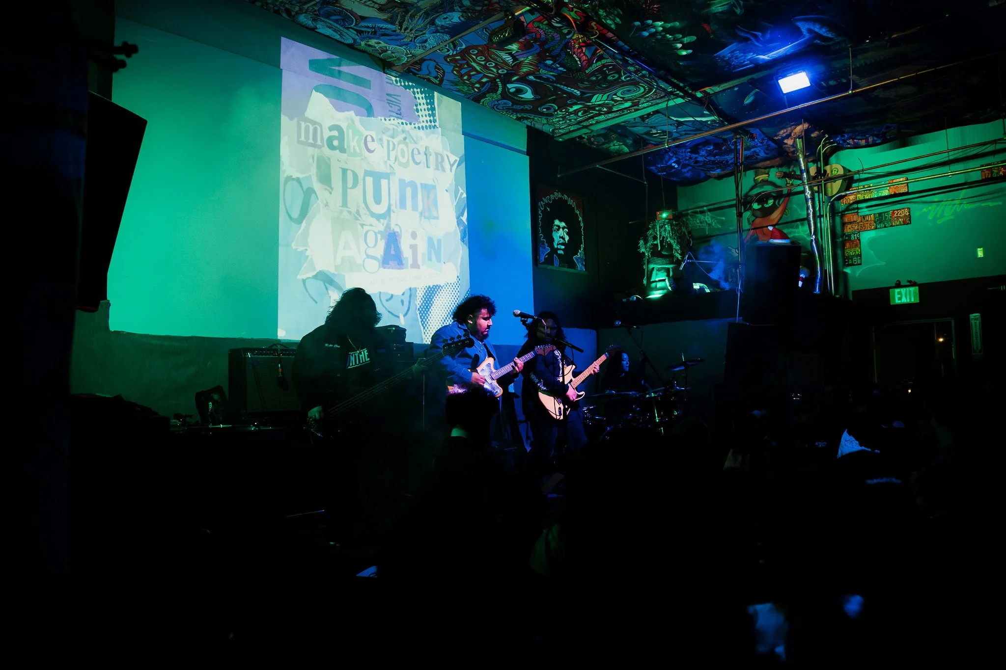 A band performing live on stage with purple and blue lighting, with a large screen displaying colorful text behind them that reads 'make poetry PUNK AGAIN'.