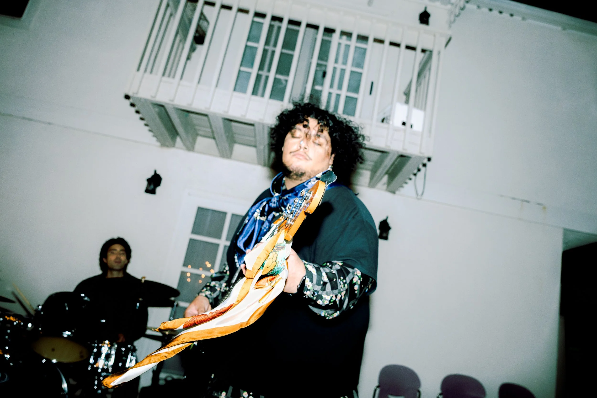 A person playing the guitar with a person on drums in the background, inside a house with a balcony above.