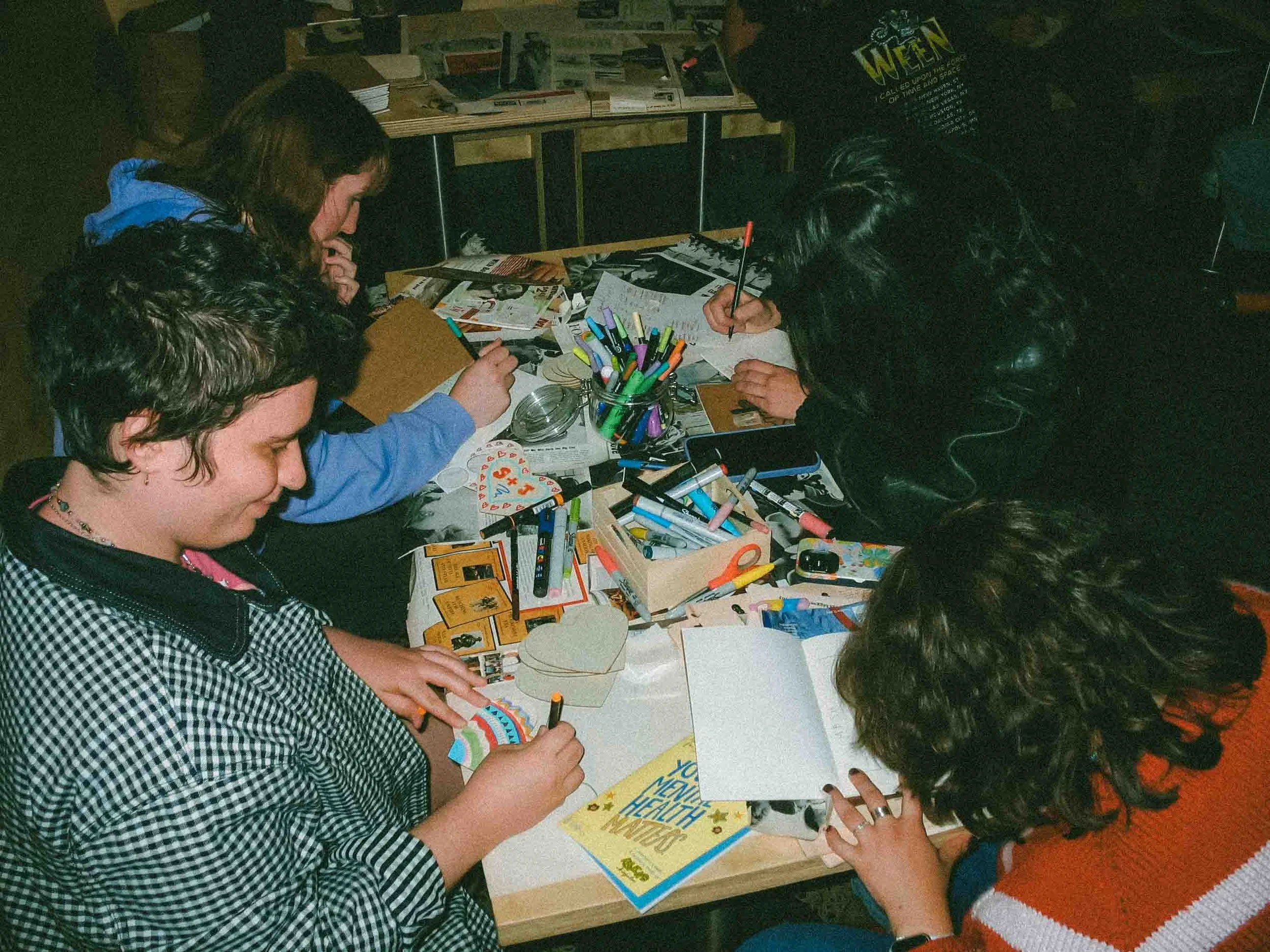 Group of people sitting around a table engaged in arts and crafts activities with various colorful markers, paper, and craft supplies.