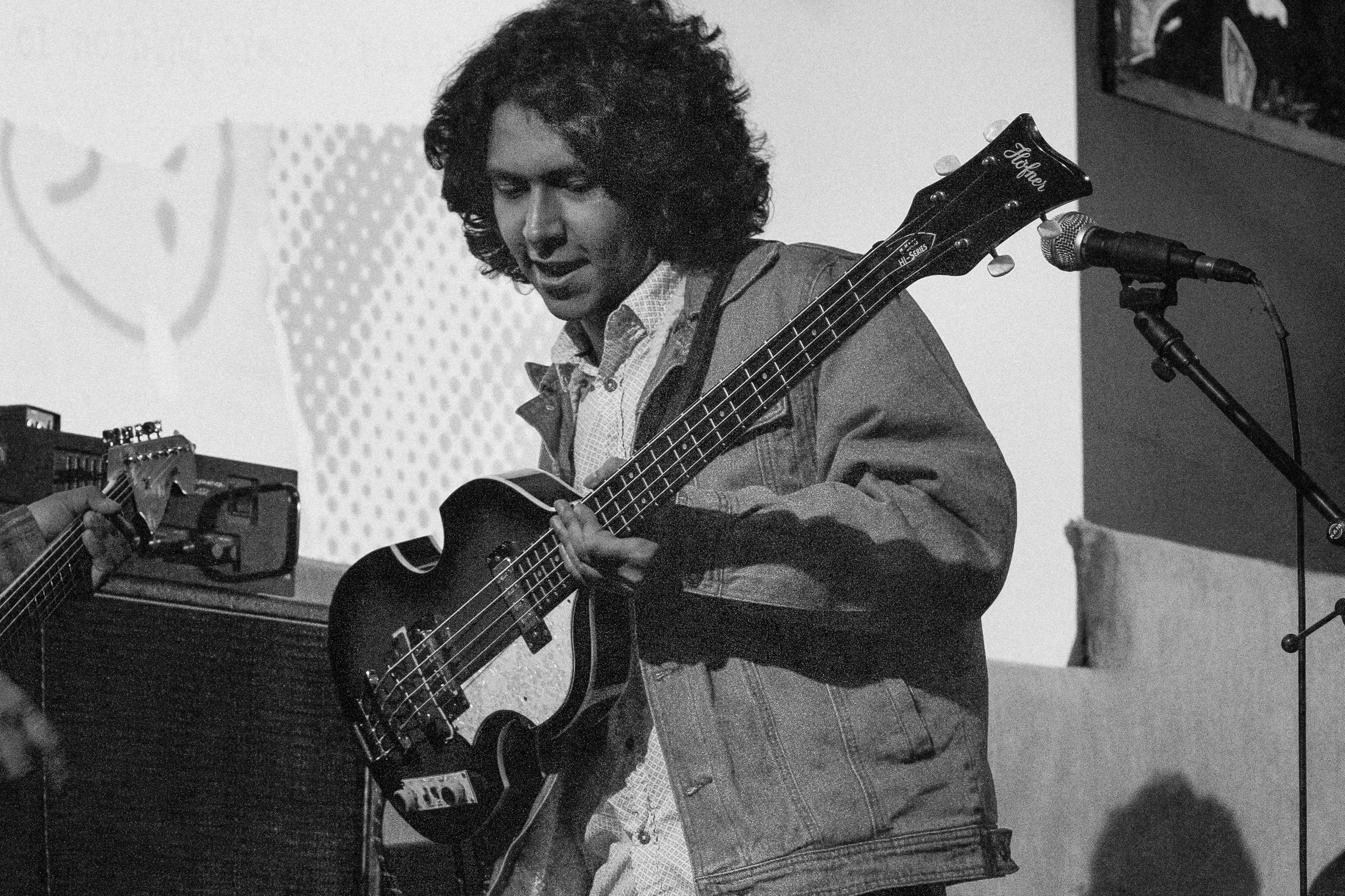 A young man with curly hair playing an electric bass guitar in a music studio, with a microphone stand nearby and a person playing a guitar partially visible on the left.