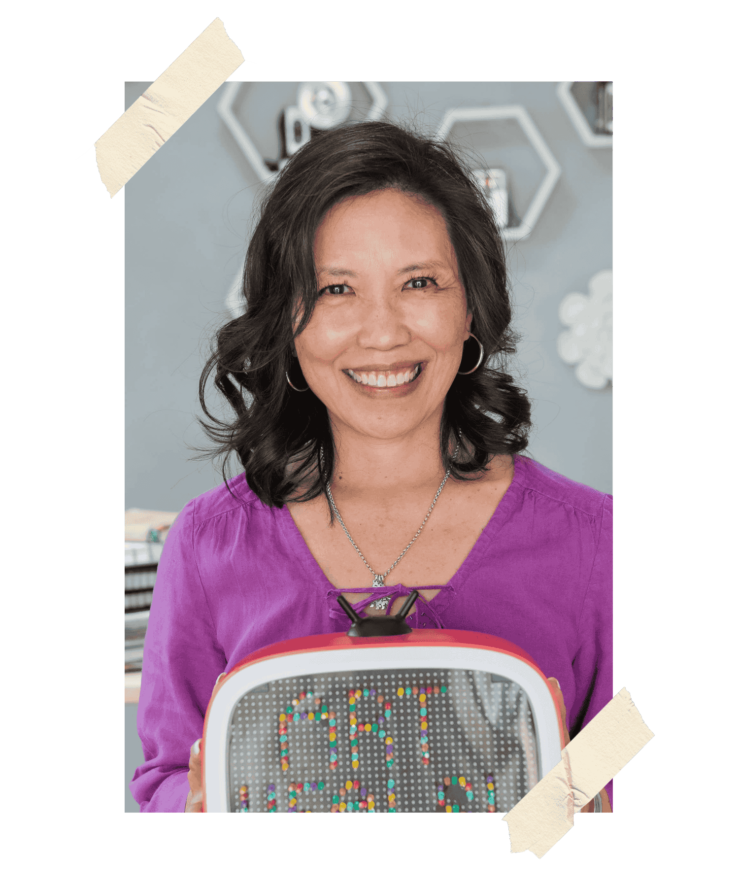 A woman with shoulder-length black hair, wearing a purple top and silver jewelry, smiling and holding a magnetic board with colorful letters that spell out 'OK!'. The background includes hexagonal shapes and a soft gray wall.