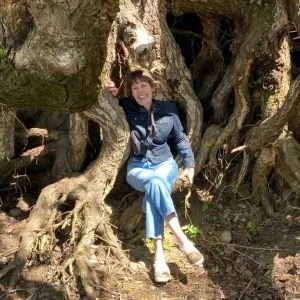 A woman with short hair wearing a black jacket and light blue pants sits on a large, gnarled tree root in a forested area.