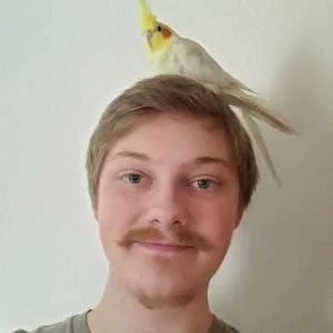 A young man with light brown hair and a mustache, smiling, has a yellow and white bird perched on his head.