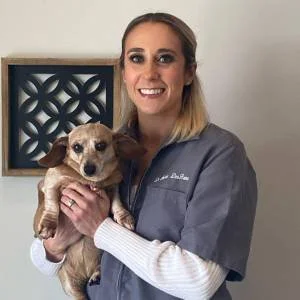 A smiling woman in scrubs holding a small tan dog in front of a decorative wall piece.