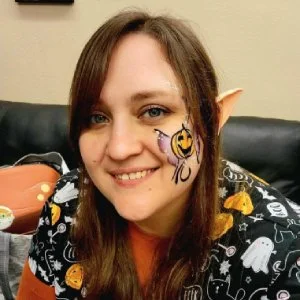 Woman with painted face of a purple spider with yellow eyes, smiling, sitting in front of a black couch with Halloween decorations.