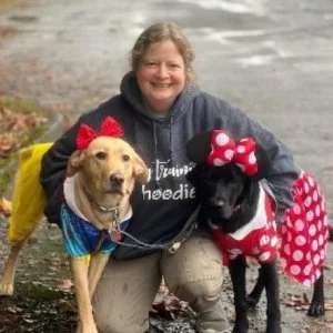 A woman smiling outdoors with two dogs dressed in costumes, one in a blue outfit with a red bow and the other in a red polka dot dress with a bow, on a rainy day by a paved path.