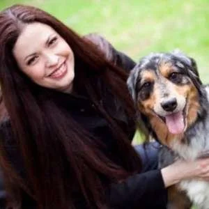 A woman smiling and sitting outdoors with a happy dog that has a multicolored coat and a pink tongue hanging out.