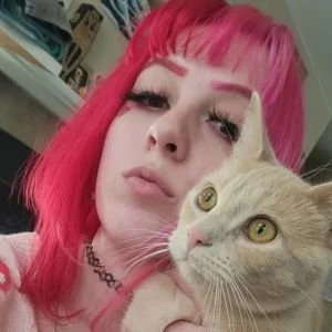 A young woman with bright pink hair and winged eyeliner taking a selfie with her beige cat.