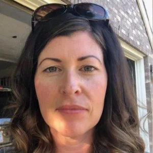 Close-up of a woman with long dark hair and sunglasses on her head, standing outdoors near a window.