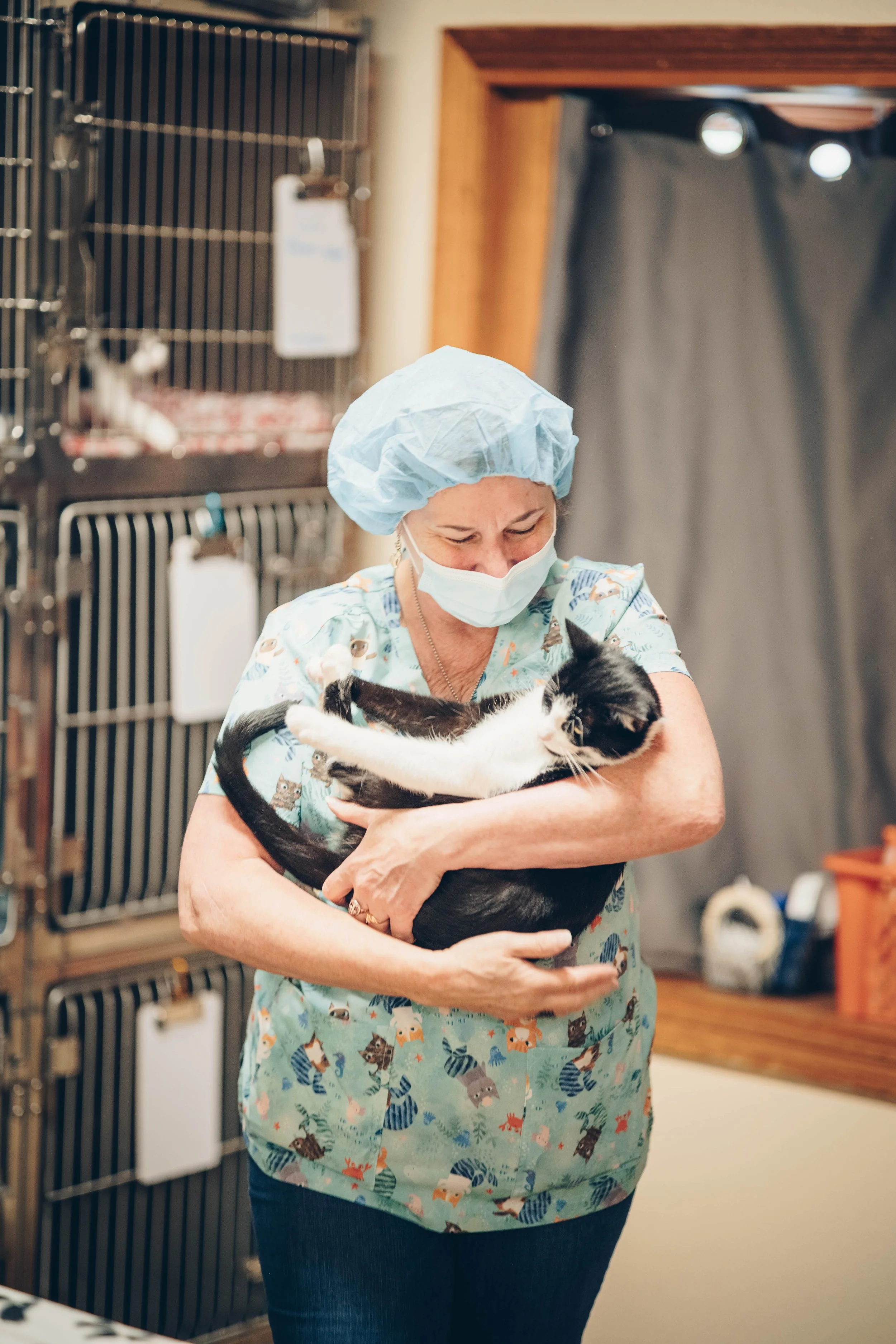 A veterinarian wearing scrubs, a face mask, and a hair cap holding a black and white cat in a veterinary clinic.