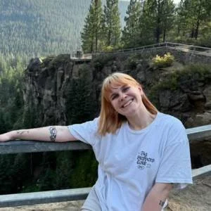 A smiling woman with blonde hair wearing a white t-shirt, standing outdoors near a railing with trees and a cliff in the background.