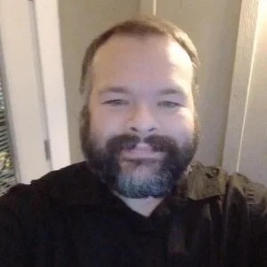 A person with a beard and mustache smiling for a selfie, wearing a dark shirt, indoors near a doorway and window.