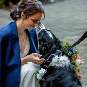 A woman with dark hair in a bun, wearing a blue jacket, interacts with two black and white dogs near water, with flowers nearby.