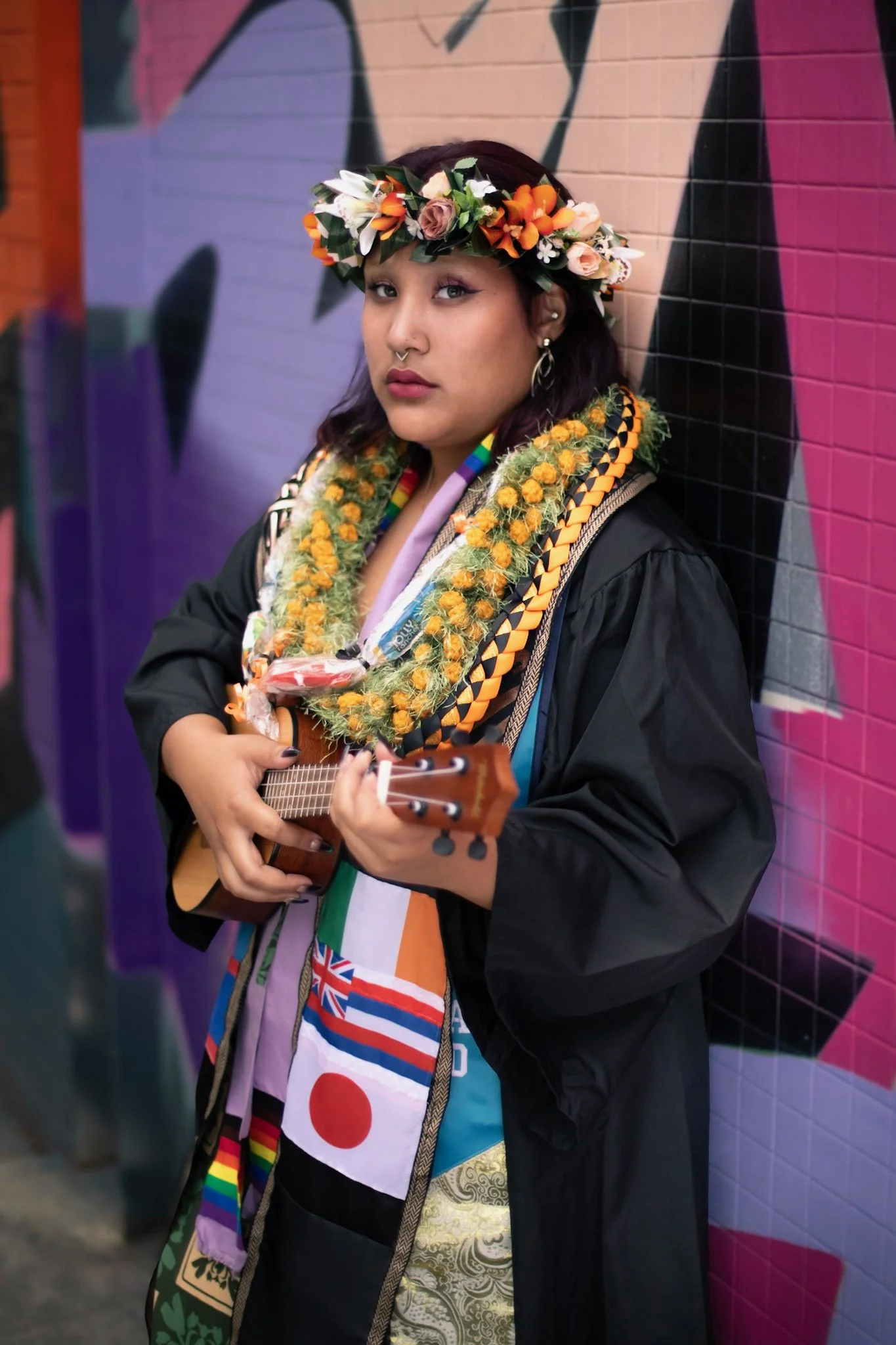 A young woman dressed in a graduation gown and stole, wearing a flower crown, holds a ukulele and stands against a colorful street mural background.