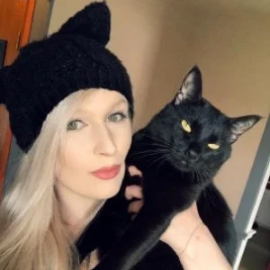 Woman with long blonde hair wearing a black knit hat, holding a black cat with yellow eyes.